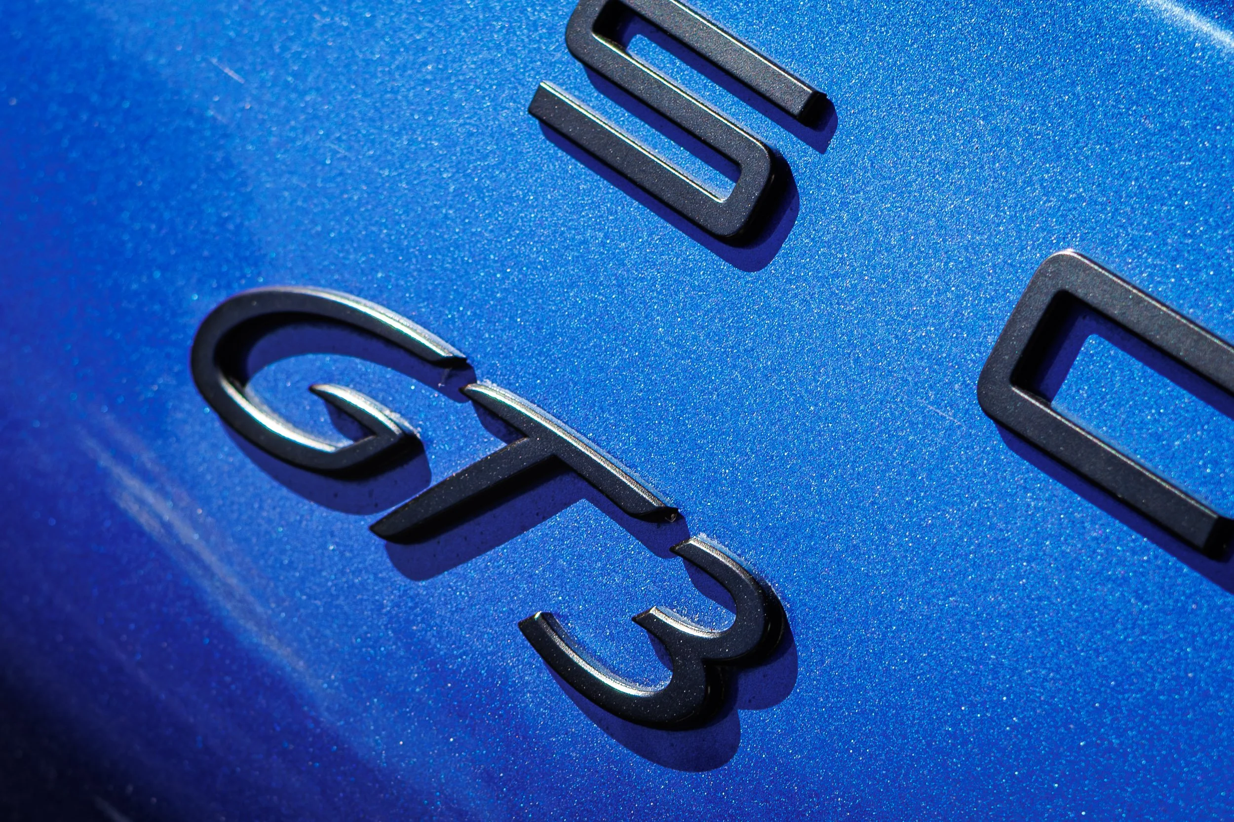 Macro detail car photography focusing on the matte black GT3 emblem and vibrant metallic blue paint flake of a Porsche 911 in Mesa, Arizona, captured by Curtis from Gasoline and Dopamine Car Photography.