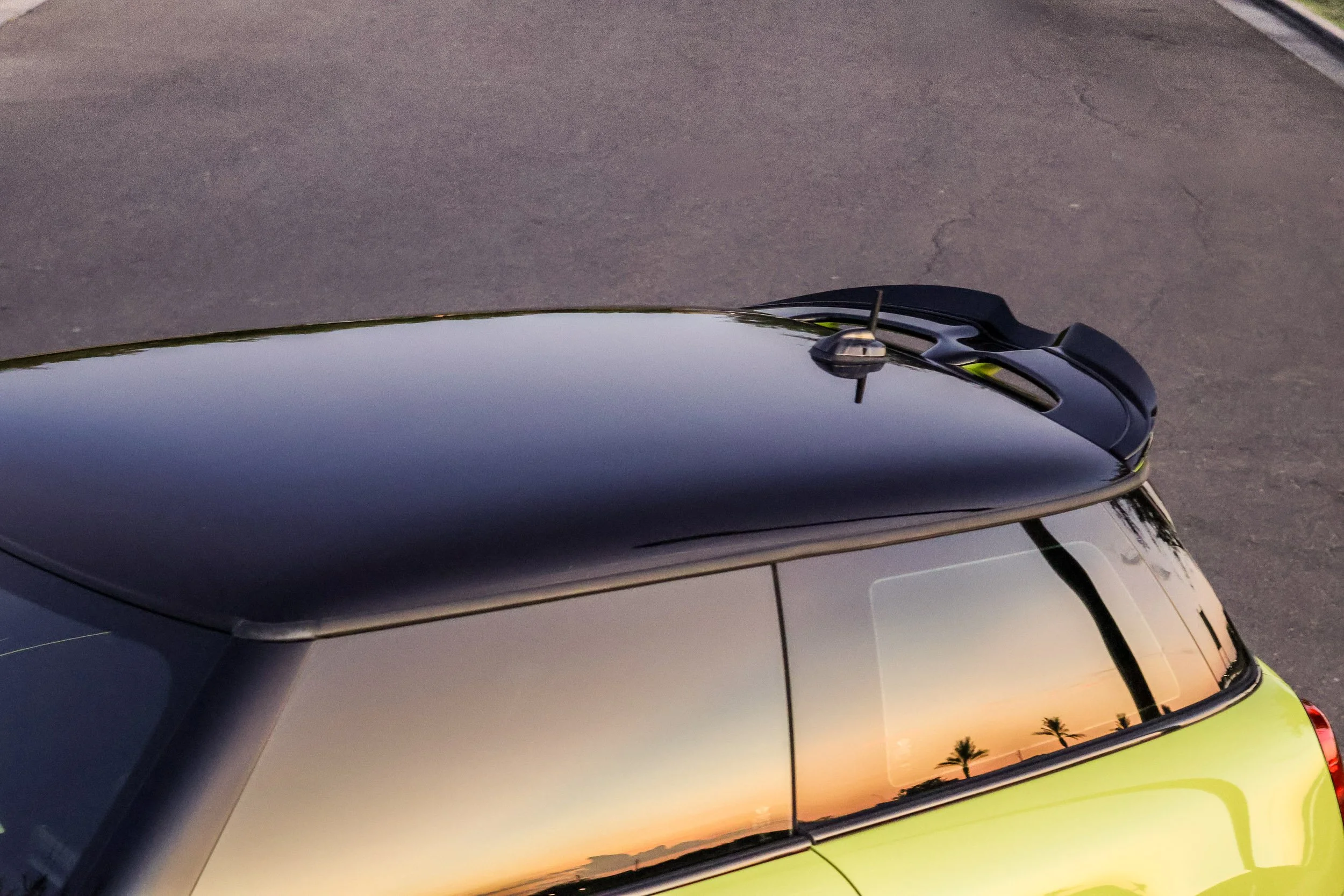 High-angle automotive photography capturing a vibrant sunset and palm tree silhouettes reflected in the tinted side glass of a yellow Mini Cooper JCW, highlighting the contrast of the black roof and rear spoiler at Morrison Ranch in Gilbert, Arizona,