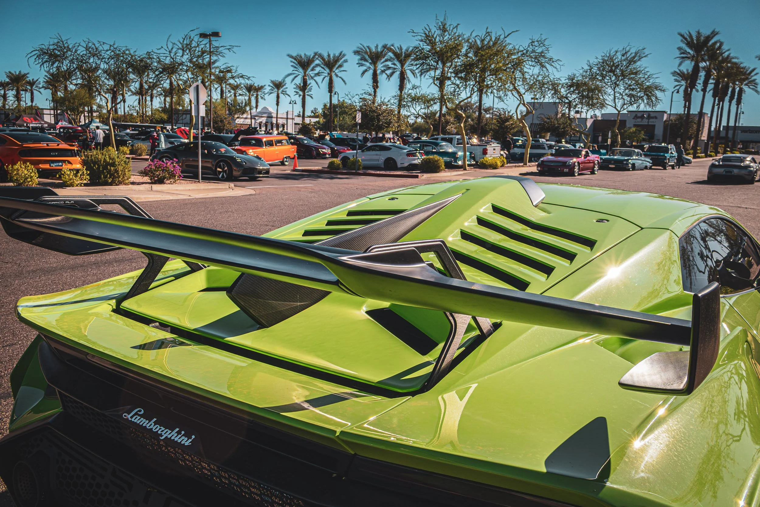 Low-angle rear automotive photography focusing on the aggressive carbon fiber rear wing, louvered engine cover, and vibrant green paint of a Lamborghini Huracan parked among a crowd of enthusiasts at a sunny, palm-lined car meet in Mesa, Arizona, cap