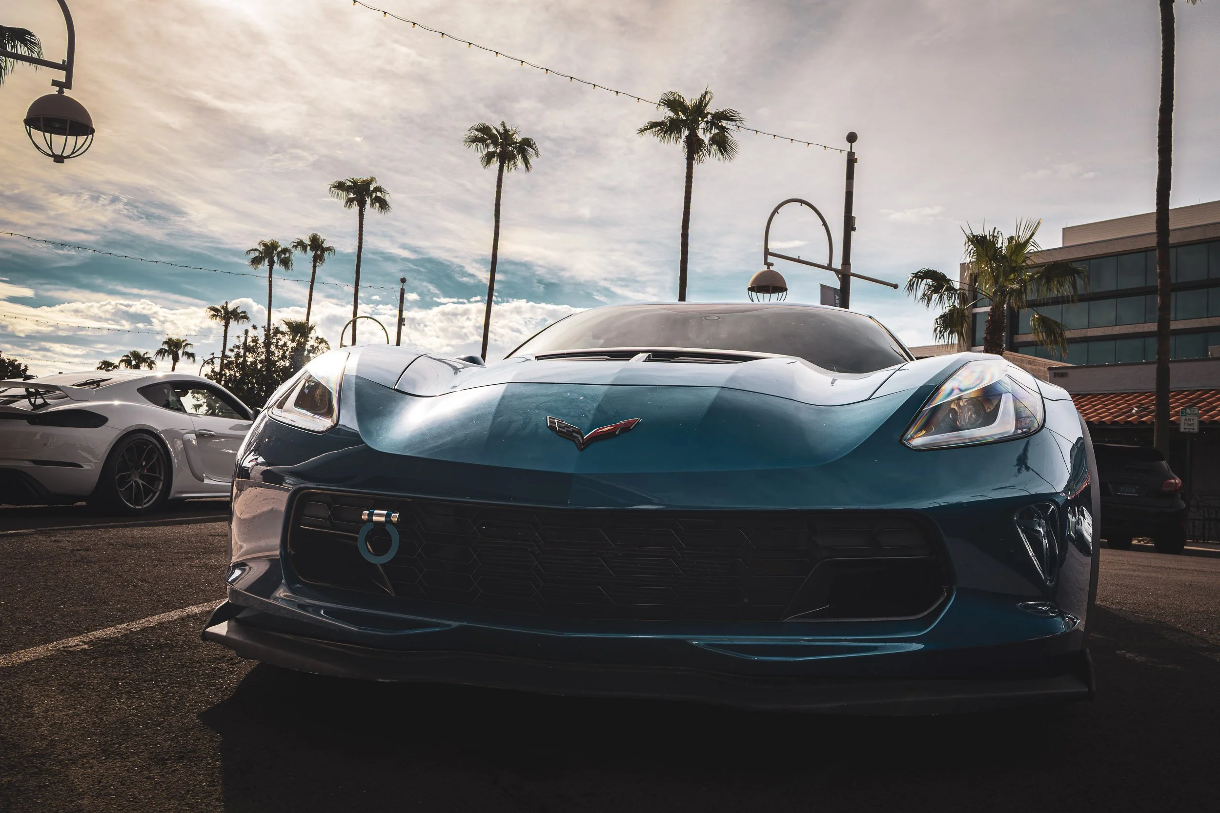Low-angle front automotive photography highlighting the aggressive front fascia, hood vent, and track-ready tow hook of a metallic blue Chevrolet Corvette C7 Z06, parked alongside a white Porsche under towering palm trees at a car meet in Scottsdale,