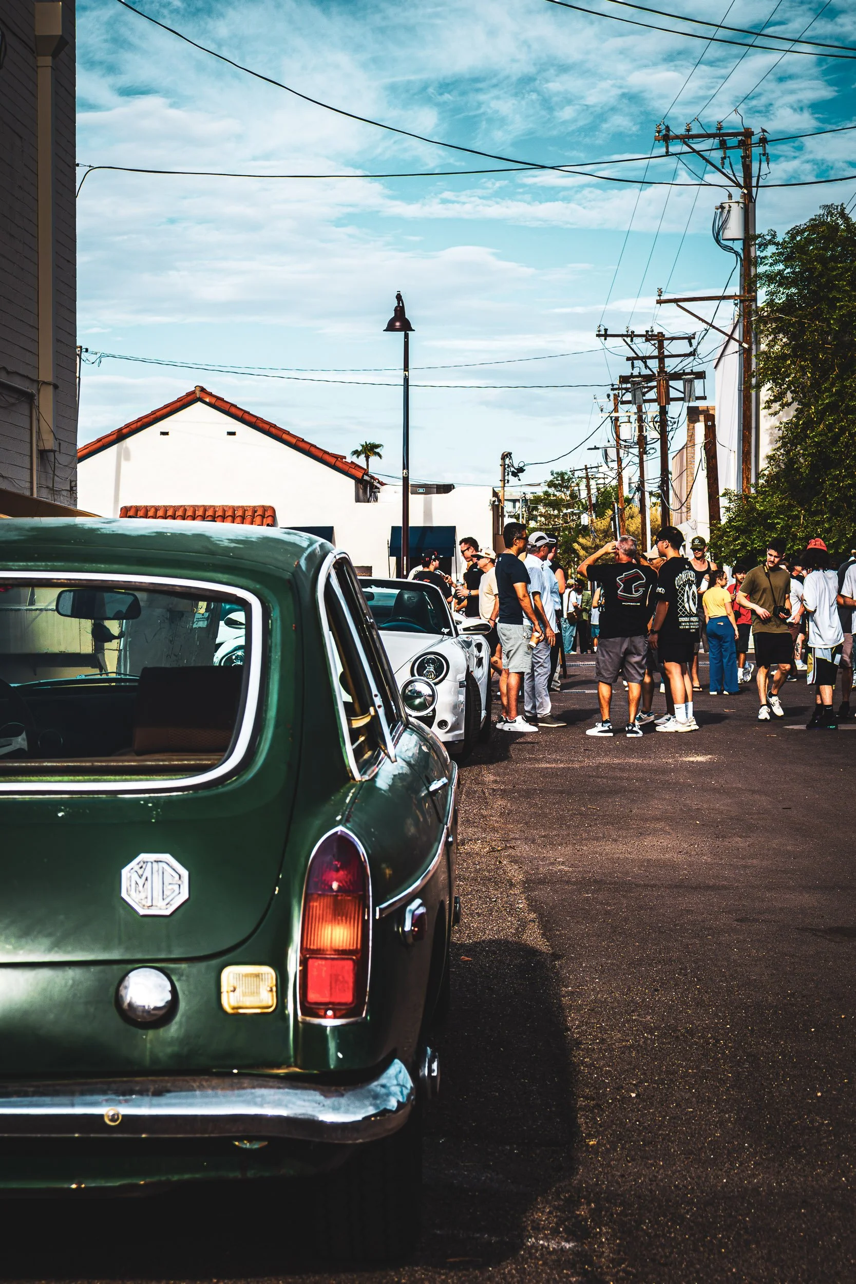 Rear profile automotive photography featuring a classic dark green MG B GT with a chrome bumper and iconic badge, parked on a street lined with automotive enthusiasts during a bustling cars and coffee event in Scottsdale, Arizona, car photography by 
