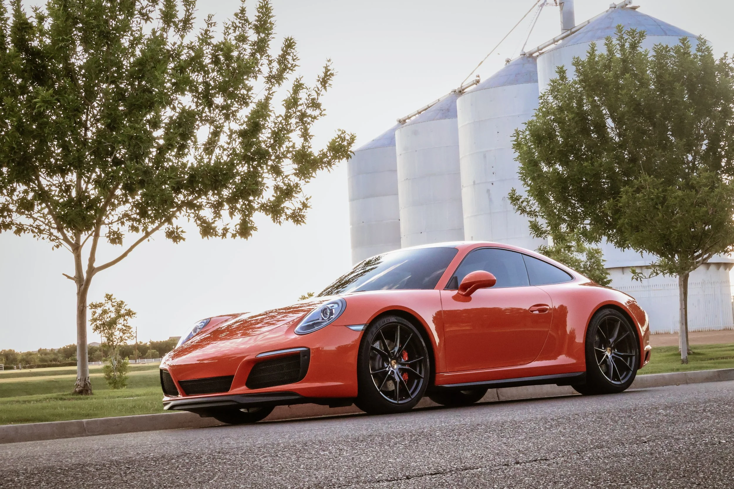 A beautiful wide shot of a 2017 Porsche 911 Carrera 4S in Lava Orange paint parked on the street under some trees with the iconic Morrison Ranch silos in Gilbert, AZ in the background.