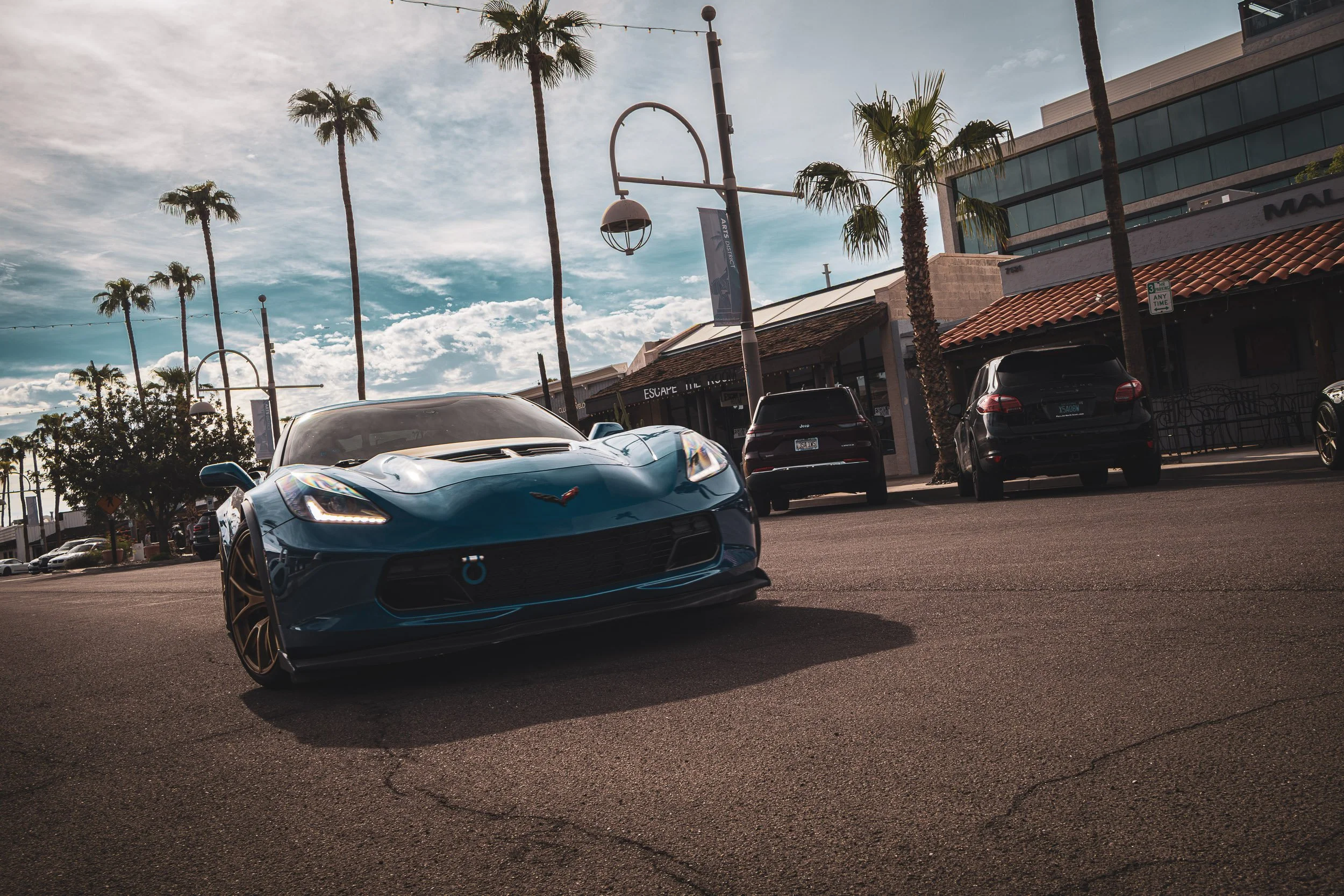 Dynamic Dutch-angle automotive photography showcasing the aggressive front fascia, illuminated headlights, and track tow hook of a metallic blue Chevrolet Corvette C7 Z06 on a sunny, palm-lined street in Scottsdale, Arizona, car photography by Gasoli