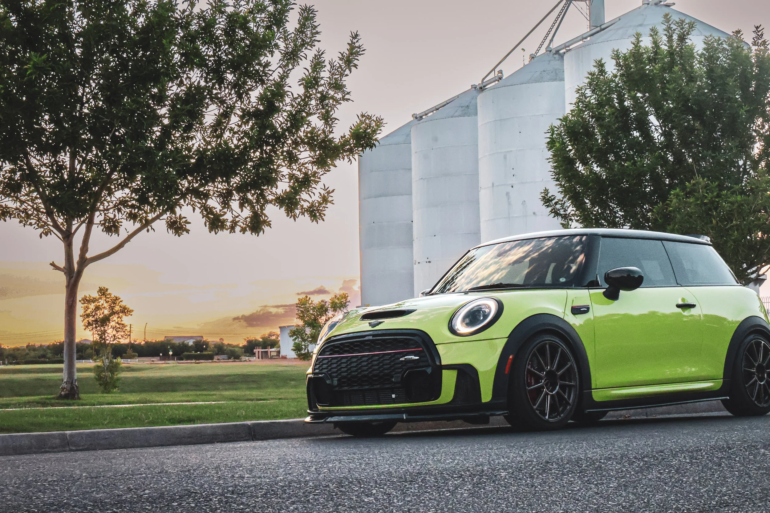 Wide-angle automotive photography showcasing a vibrant yellow Mini Cooper JCW parked along a curb during a picturesque sunset, framed by neighborhood trees and the historic Morrison Ranch silos in Gilbert, Arizona, photoshoot by Curtis at Gasoline an