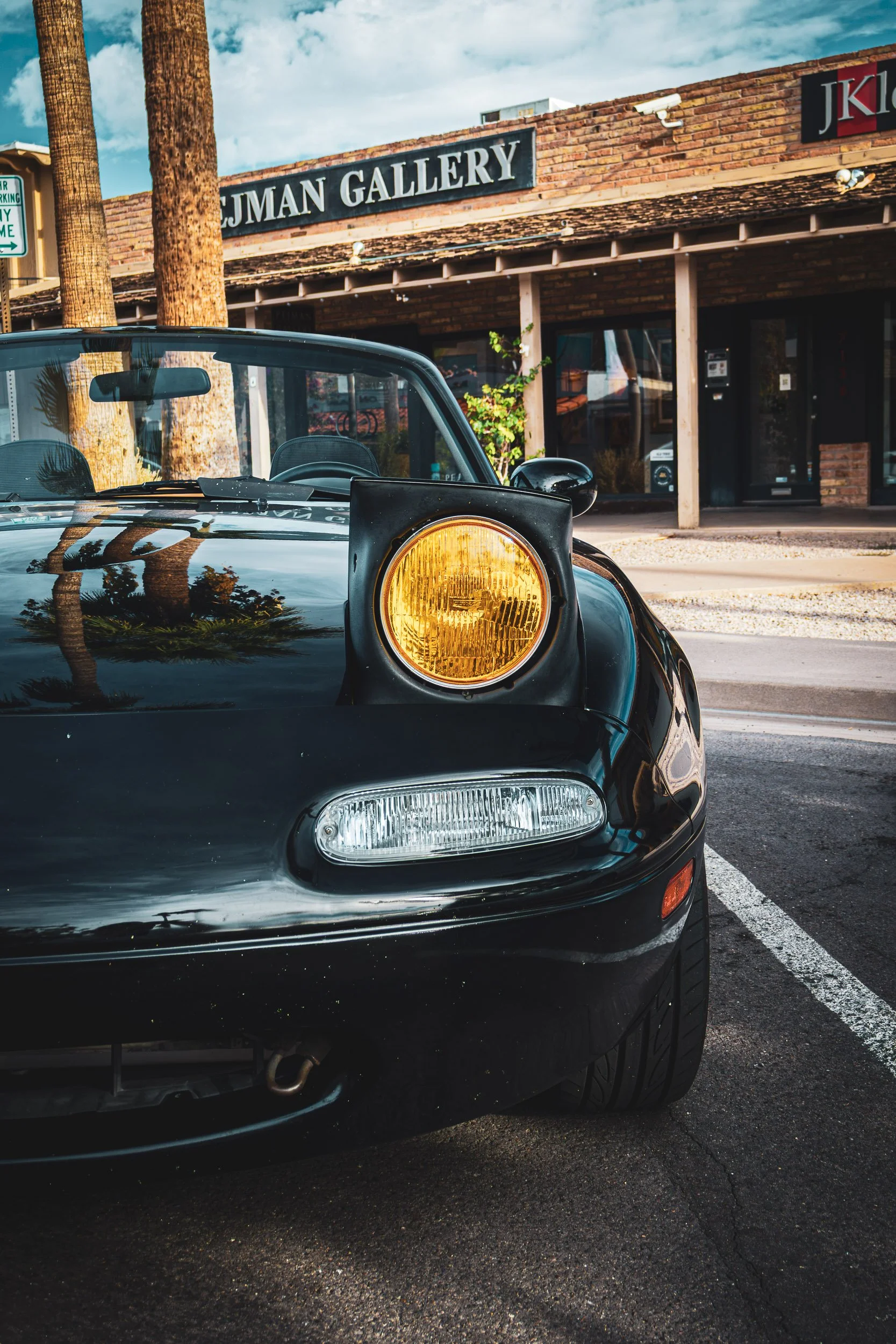 Front-facing automotive photography focusing on a classic black Mazda Miata NA featuring a raised, yellow-tinted pop-up headlight. Palm trees are reflected clearly on the glossy hood as the car sits parked on a street in front of a brick gallery buil