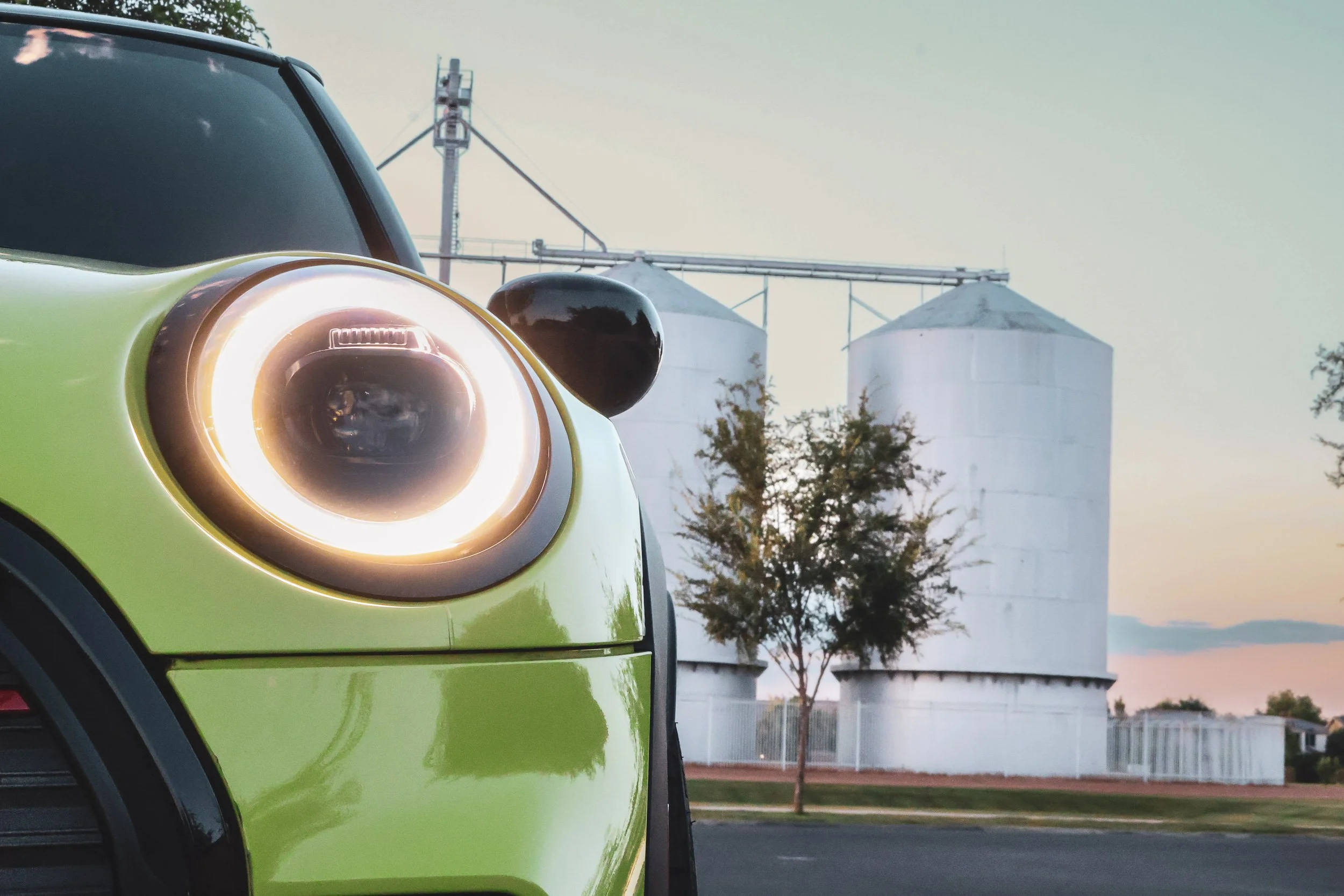 Macro automotive photography focusing on the brightly illuminated halo headlight of a yellow Mini Cooper JCW, set against the historic Morrison Ranch silos at dusk in Gilbert, Arizona, photoshoot by Curtis from Gasoline and Dopamine.