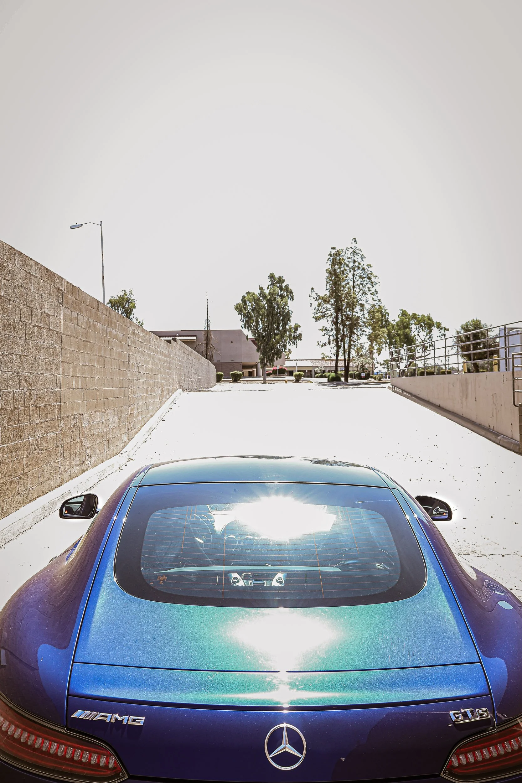 Vertical automotive photography capturing the rear profile and wide haunches of a metallic blue Mercedes-AMG GT S, parked on an industrial concrete ramp under harsh, blown-out midday sunlight in Gilbert, Arizona, photograph by Gasoline and Dopamine C