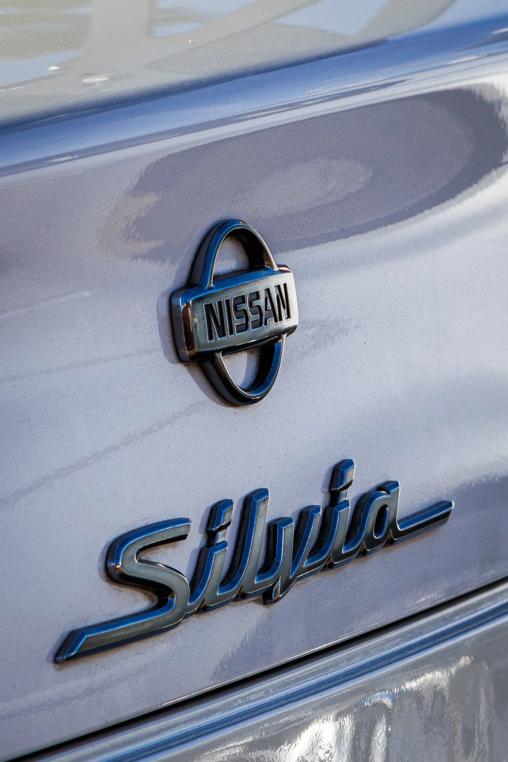 Macro detail car photography focusing on the dark chrome Nissan Silvia emblems set against silver metallic paint in Gilbert, Arizona, photo by Curtis from Gasoline and Dopamine.