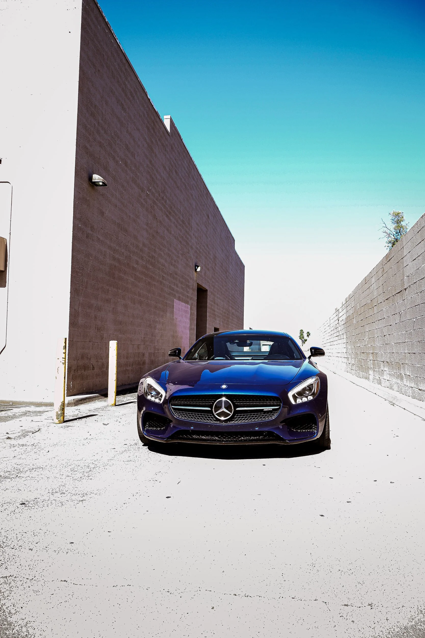 Vertical automotive photography capturing the front fascia and wide stance of a metallic blue Mercedes-AMG GT S, parked in a textured industrial alleyway under a clear blue sky and harsh midday sunlight in Queen Creek, Arizona, photo by Curtis at Gas