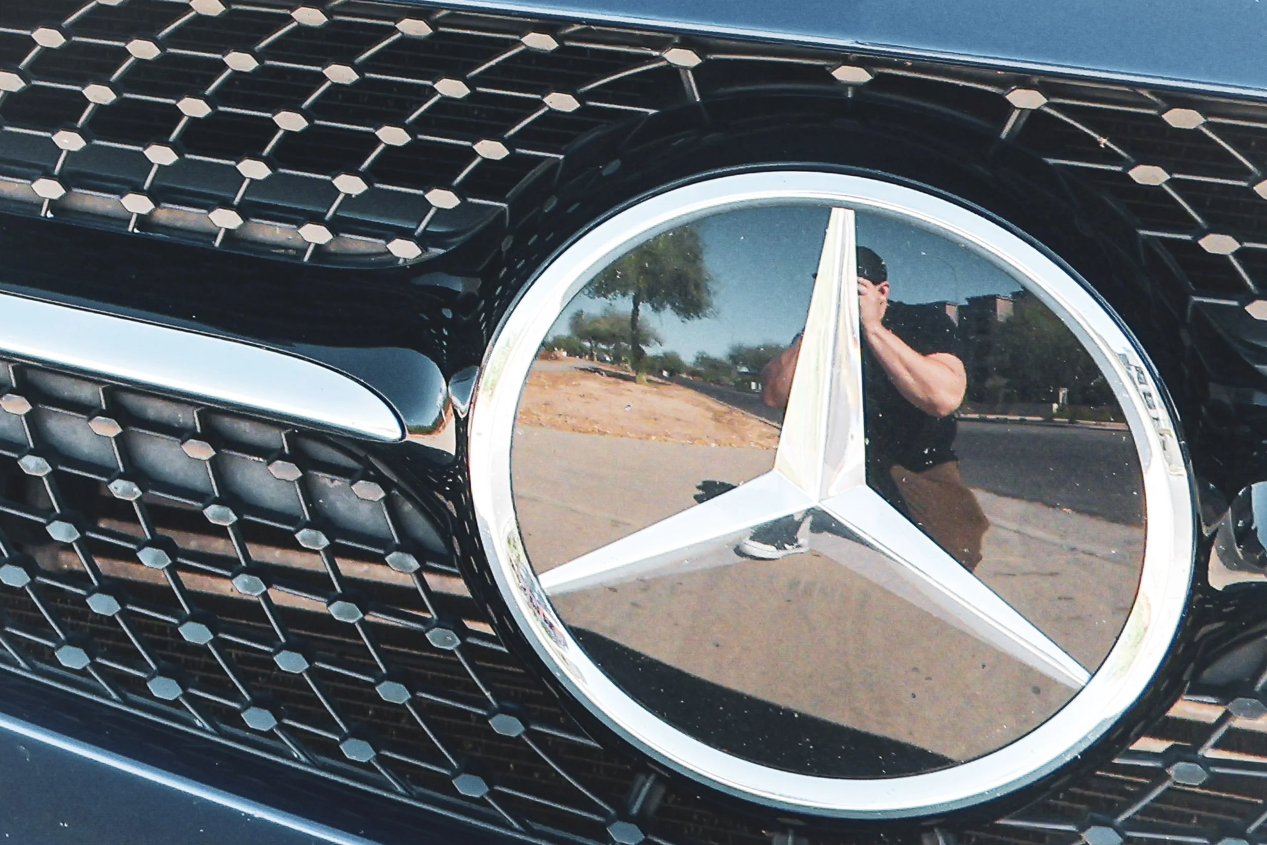 Macro automotive detail photography focusing on the iconic chrome Mercedes-Benz Tristar emblem and diamond grille of a blue Mercedes-AMG GT S, capturing a self-portrait reflection of the photographer in the badge on a sunny day in Gilbert, Arizona, p