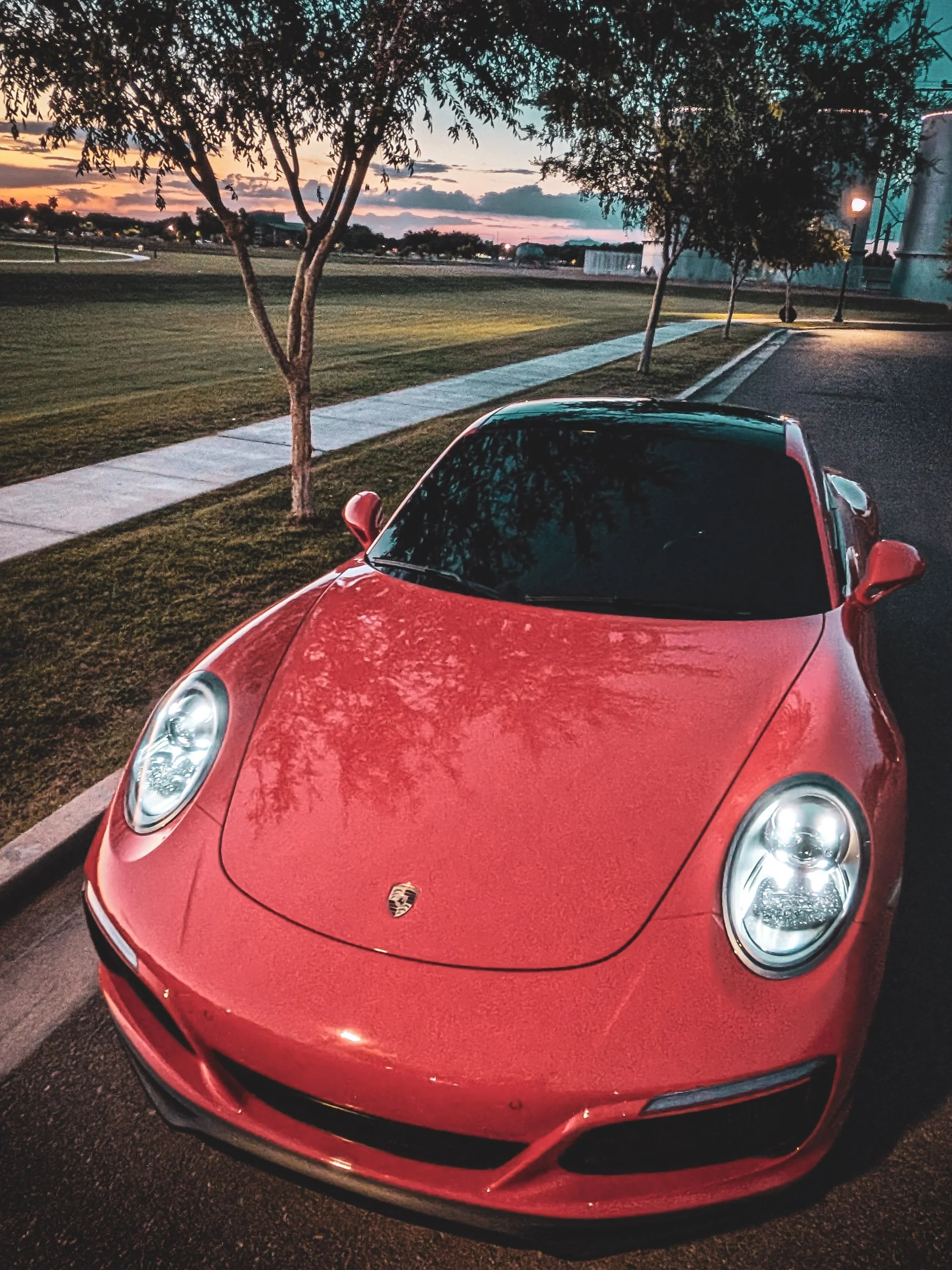 Twilight car photography capturing a high-angle view of a Lava Orange Porsche 911 C4S with illuminated headlights, parked near the Morrison Ranch silos in Gilbert, Arizona, captured by Curtis of Gasoline and Dopamine.