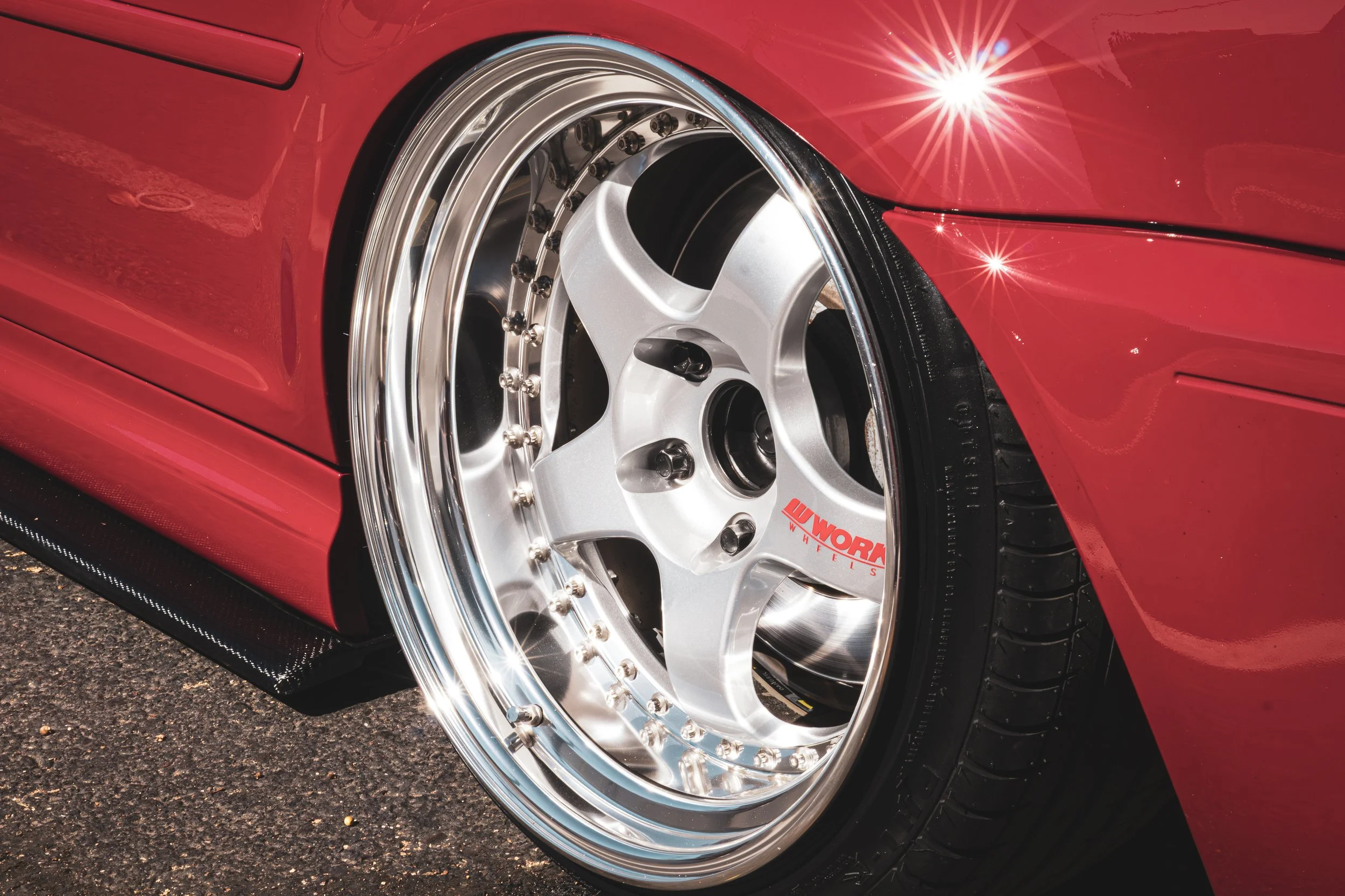 A crisp 50mm close-up shot of a beautiful silver 5-spoke Works wheel on a lowered old red BMW 3 series.