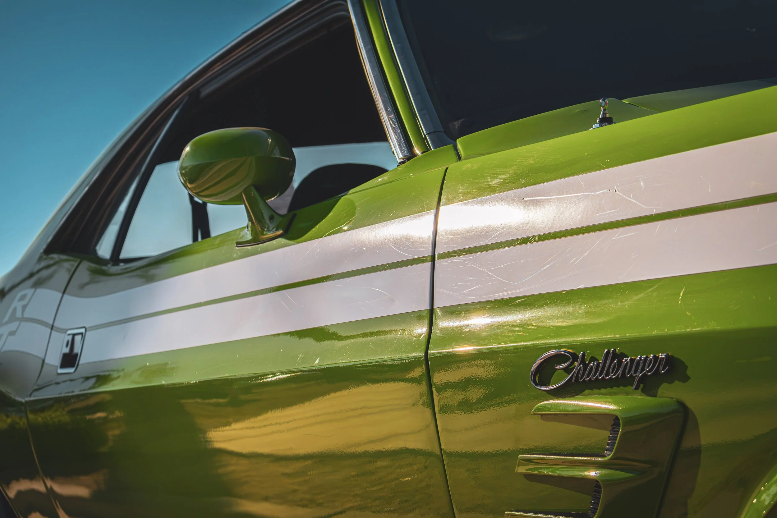 Close-up side profile automotive photography focusing on the vibrant green paint, dual white racing stripes, and classic script badge of a vintage Dodge Challenger R/T reflecting the bright Mesa, Arizona sunlight, captured by Curtis for Gasoline and 