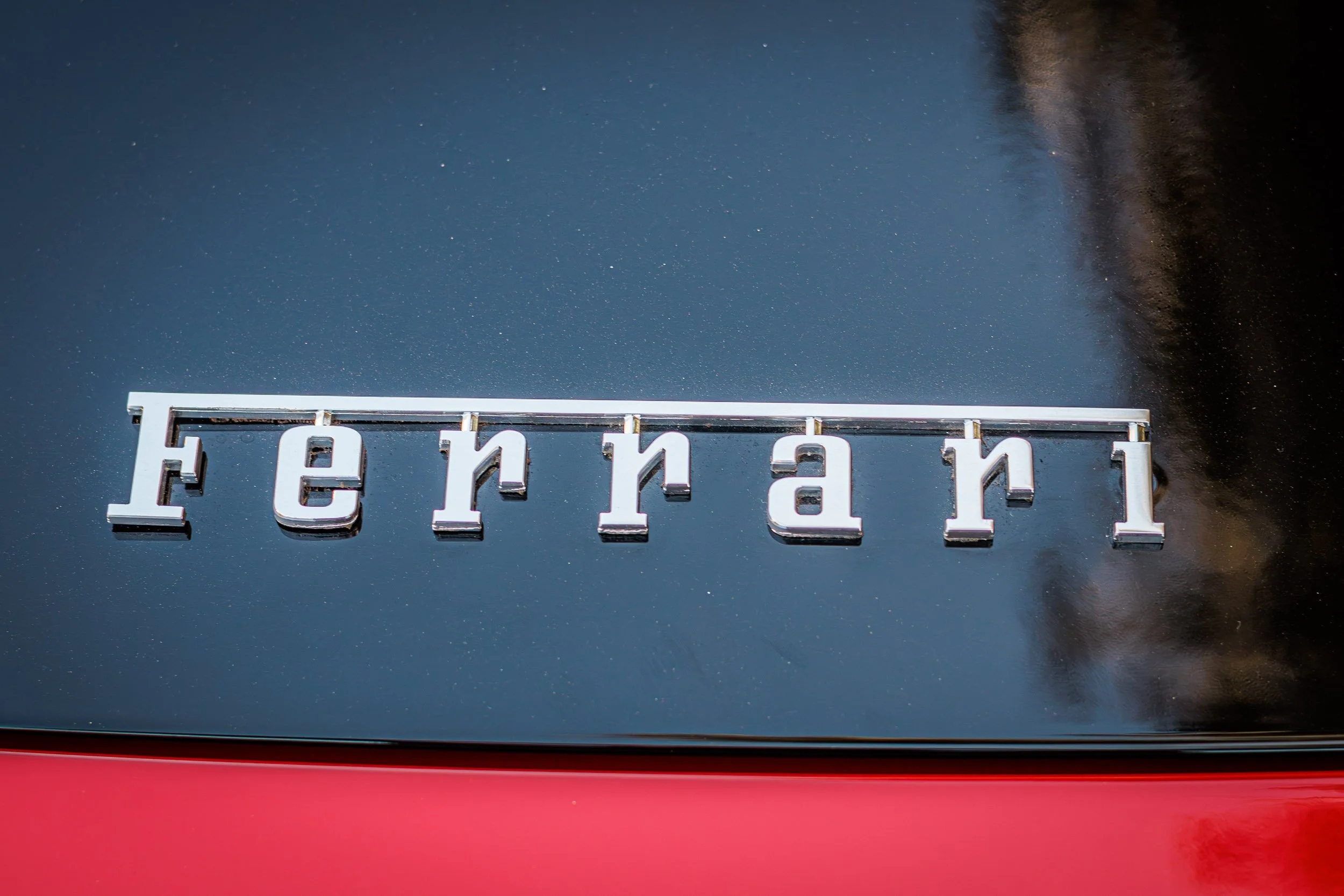 Macro detail exotic car photography highlighting the iconic chrome Ferrari script emblem resting on a glossy black rear decklid with a vibrant red lower panel, captured by Curtis from Gasoline and Dopamine in Gilbert, Arizona.