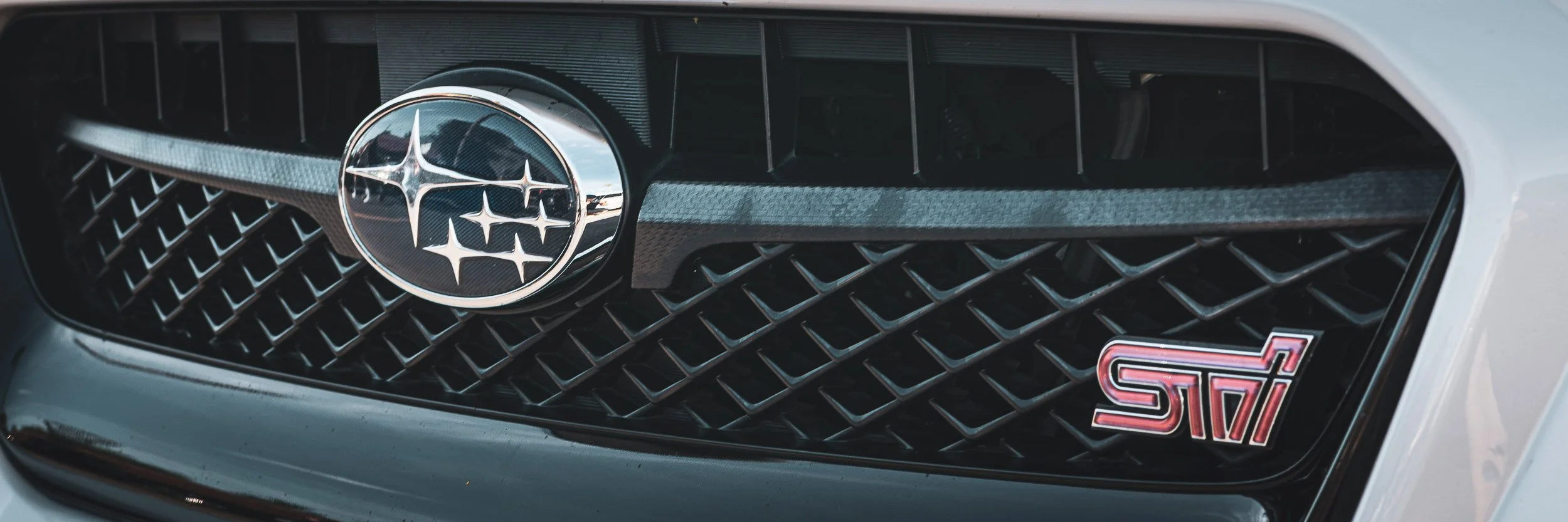 Macro detail automotive photography focusing on the iconic chrome Subaru star cluster emblem and red STI badge mounted on the black mesh front grille of a Subaru WRX STI in Arizona, captured by Curtis from Gasoline and Dopamine.