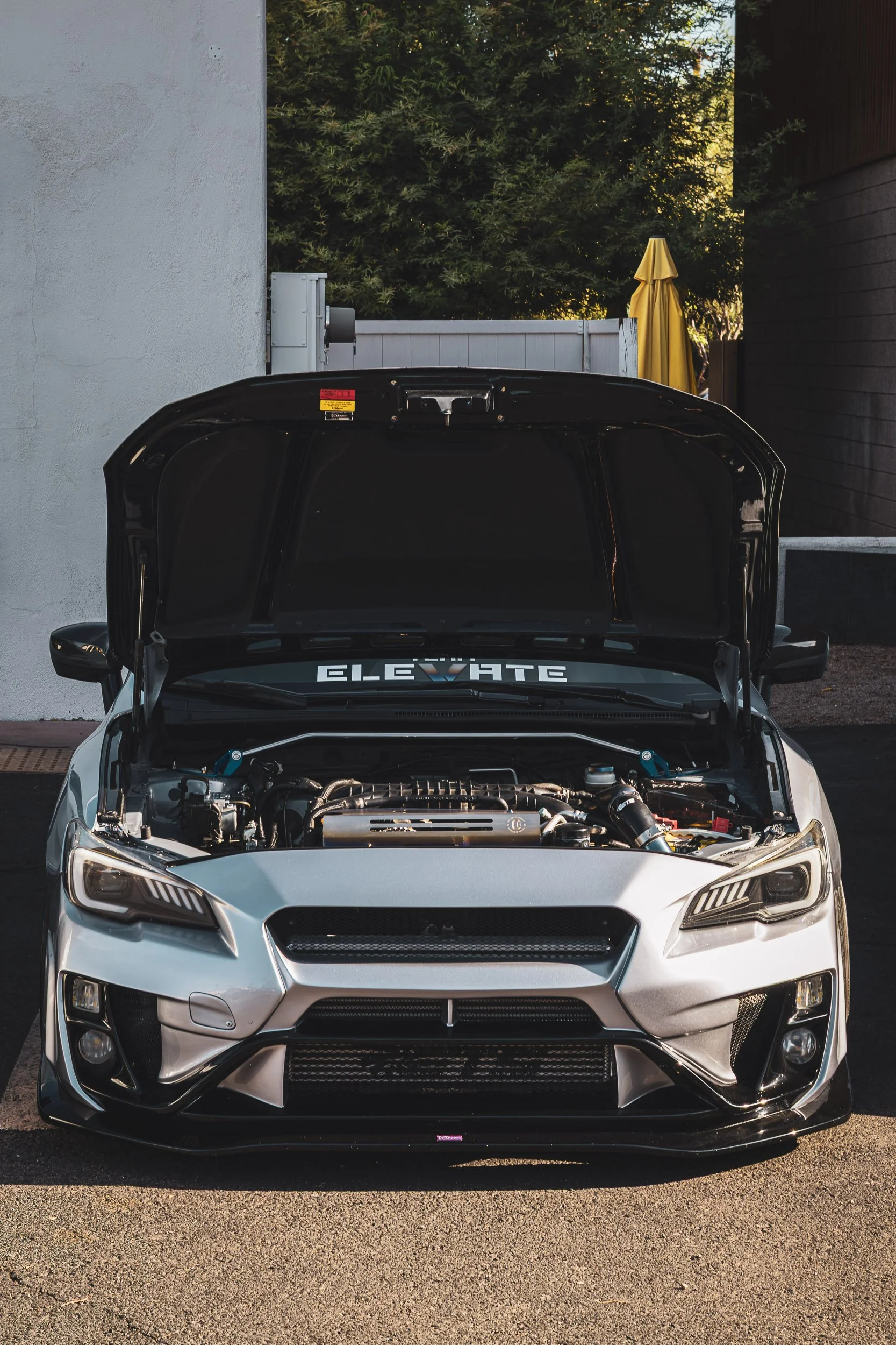 Vertical front-facing automotive photography showcasing a modified silver Subaru WRX with an open hood, exposed engine bay, aggressive front aero, and an "ELEVATE" windshield banner, captured by Curtis at Gasoline and Dopamine in Scottsdale, Arizona.