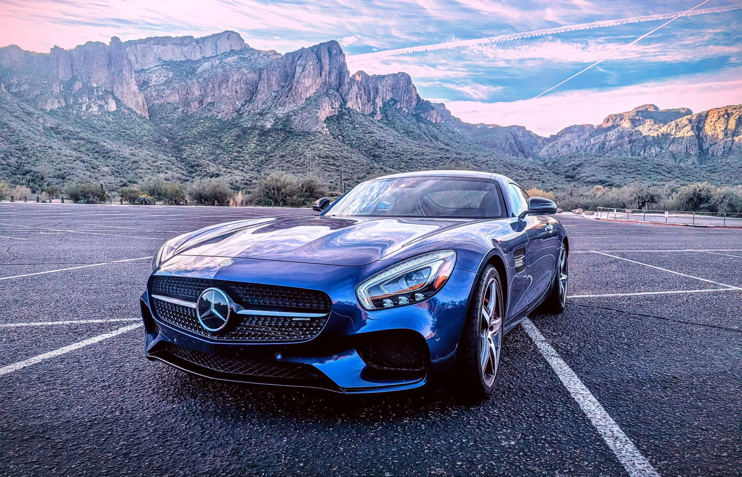 Low-angle front three-quarter automotive photography featuring a metallic blue Mercedes-AMG GT S coupe parked in an empty lot, set against the rugged Superstition Mountains under a vibrant sunrise sky in Arizona, photo captured by Curtis from Gasolin