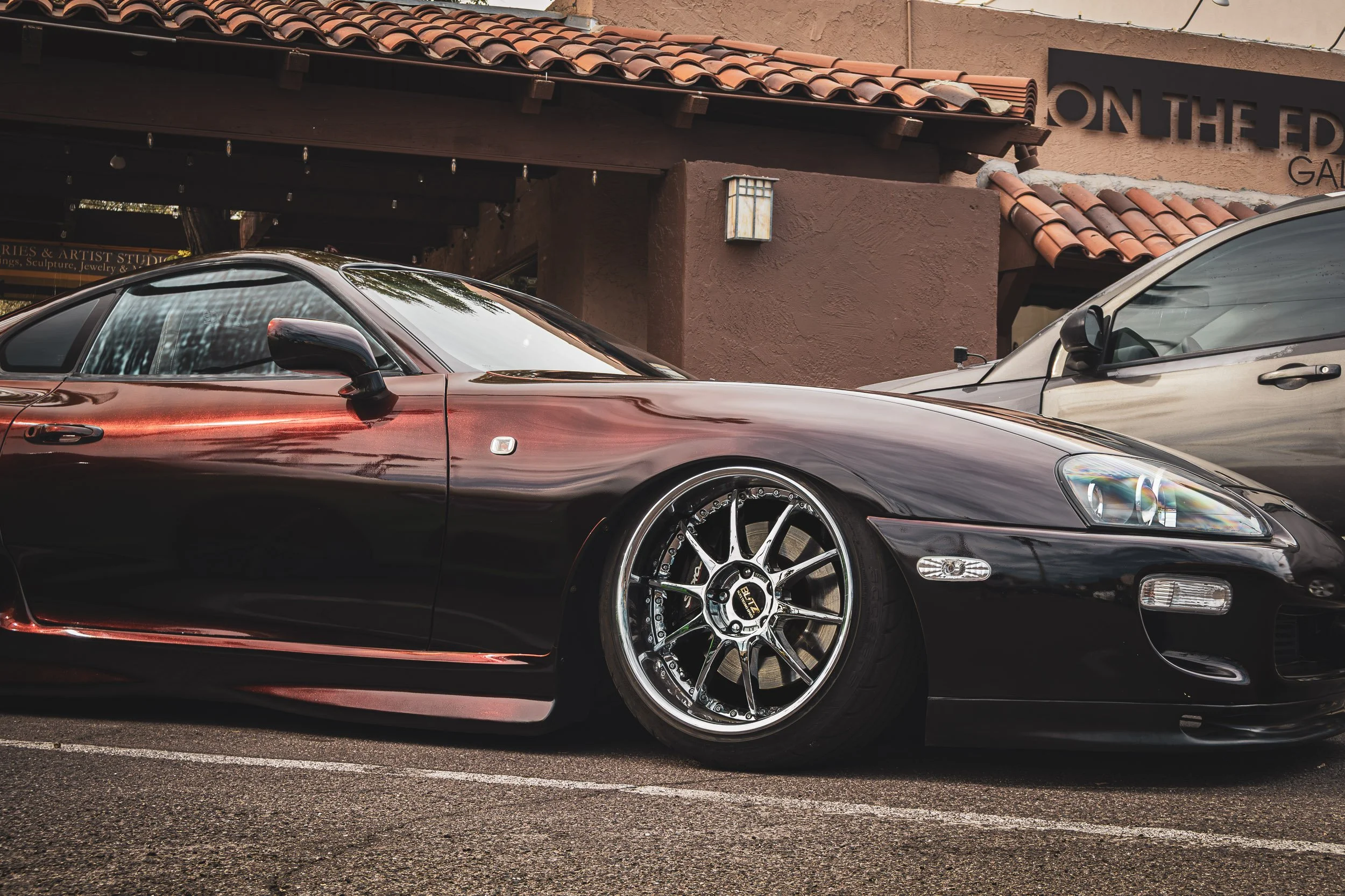 Low-angle front three-quarter automotive photography showcasing a modified, stanced Toyota Supra MK4 featuring a unique colorshift paint finish, deep-dish chrome BLITZ wheels, and a rear wing, parked on a street in front of an art gallery during a ca