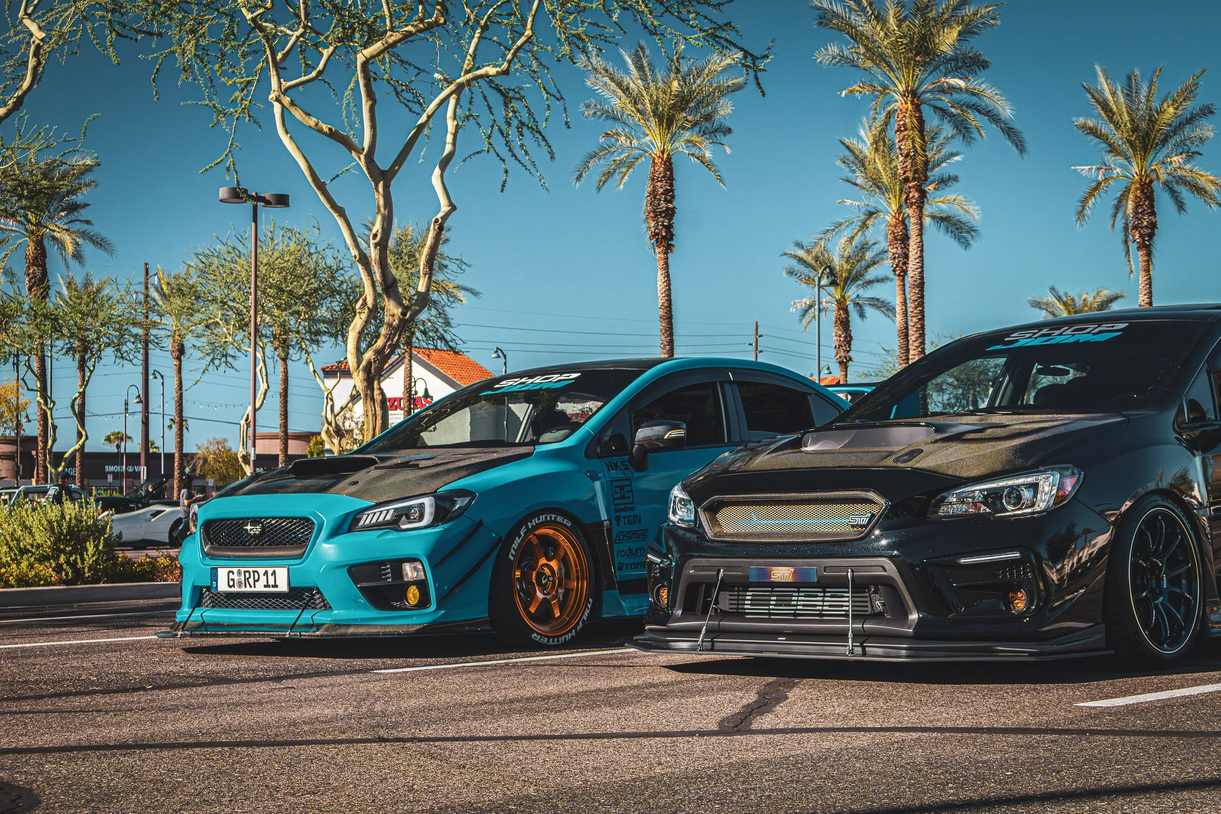 Low-angle street car photography featuring a heavily modified teal Subaru WRX and a black Subaru STI parked side-by-side, highlighting their carbon fiber hoods, aftermarket wheels, and aggressive aero under sunny skies and palm trees at a Mesa, Arizo