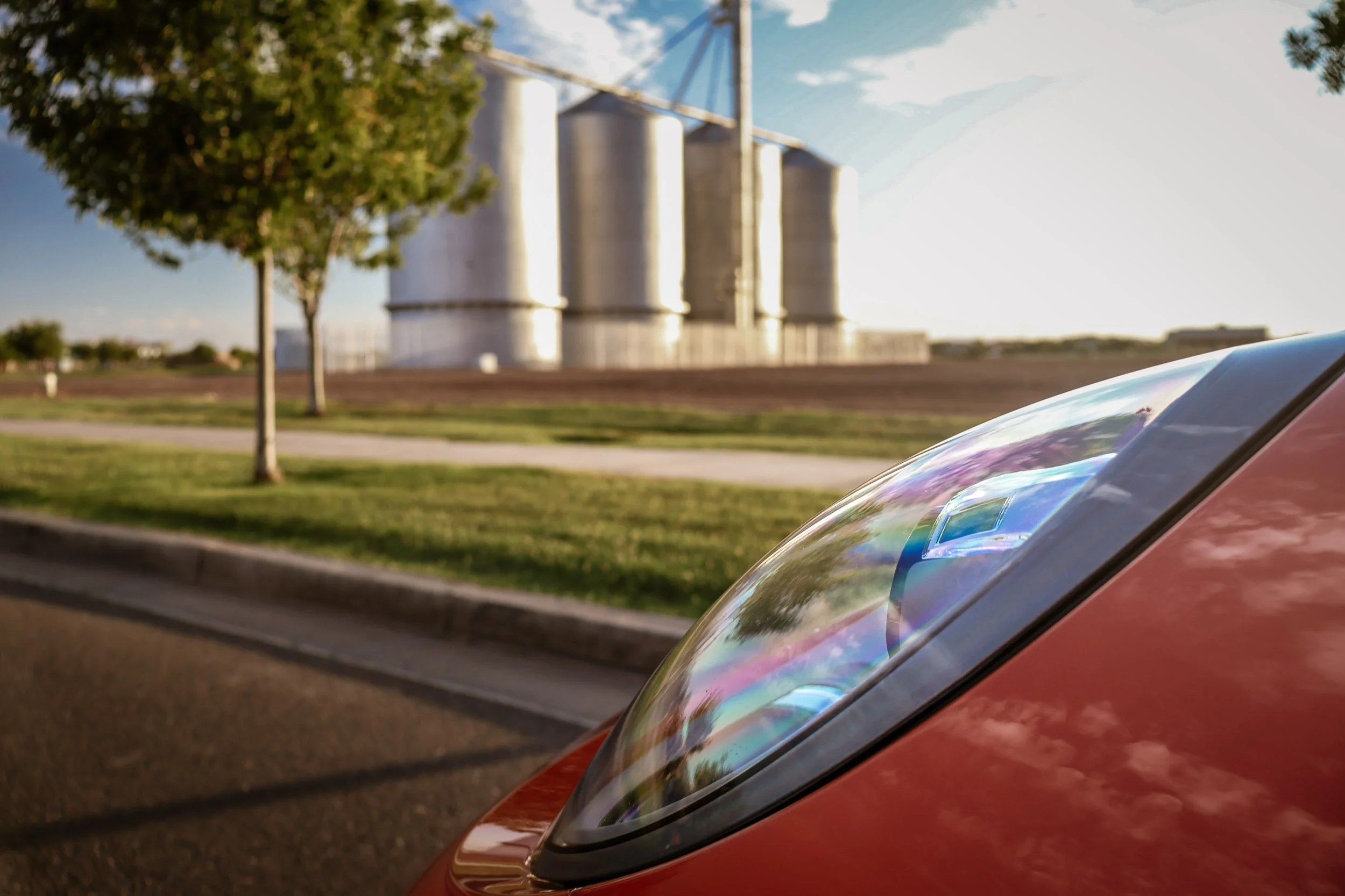 Macro side profile automotive photography focusing on the curved glass headlight of a Lava Orange Porsche 911 C4S, with the historic Morrison Ranch silos blurred in the background in Gilbert, Arizona, captured by Gasoline and Dopamine Photography.