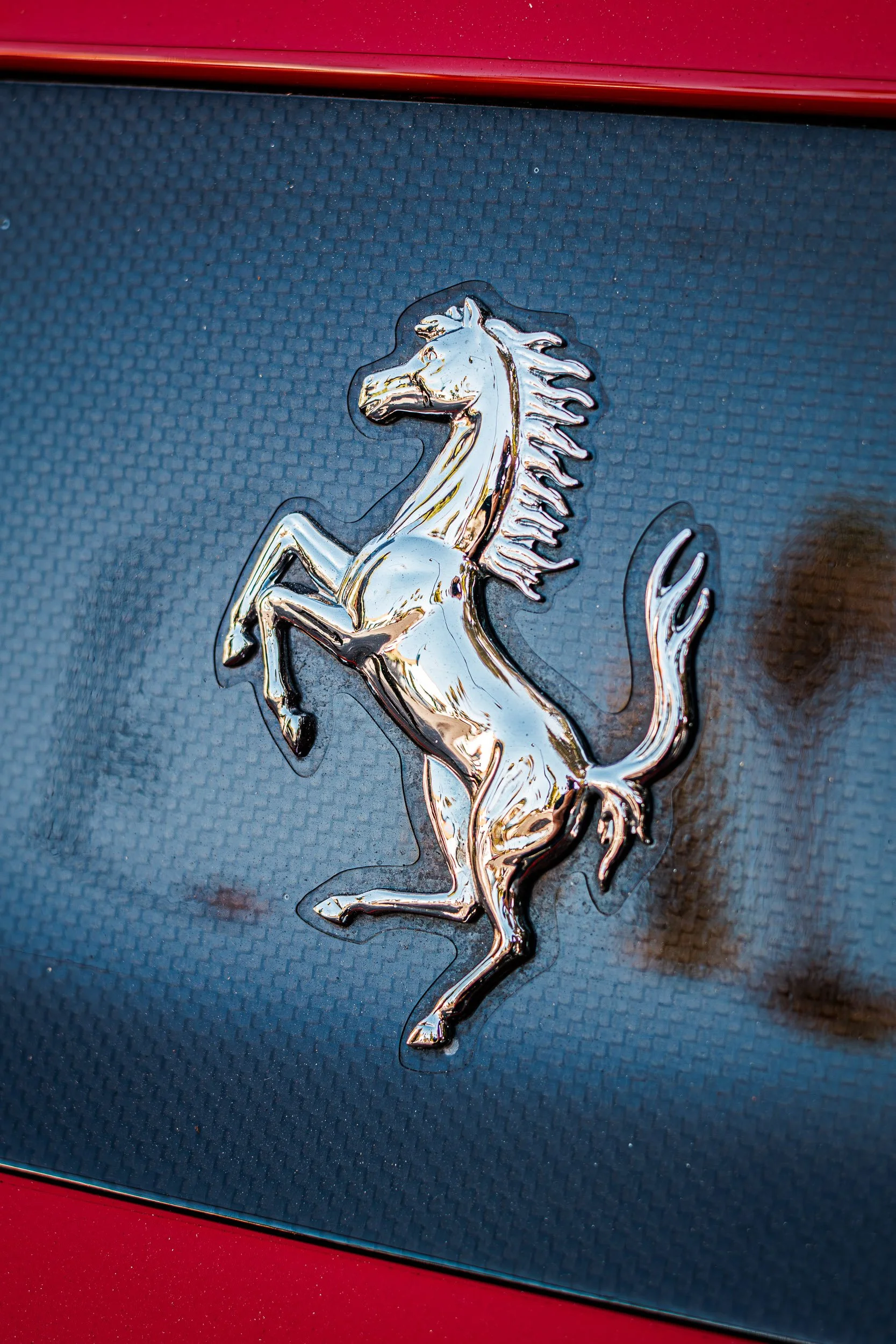 Macro detail car photography focusing on a chrome Ferrari Prancing Horse emblem mounted against a carbon fiber texture and red paint in Queen Creek, Arizona, captured by Gasoline and Dopamine.