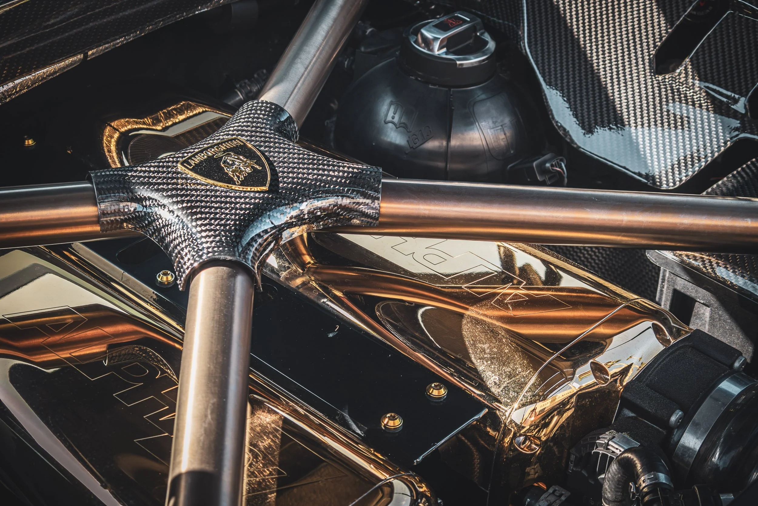 Macro detail automotive photography showcasing a gold mirror-polished AMS Performance Alpha twin-turbo engine bay, carbon fiber cross-brace with the Lamborghini crest, and titanium support bars on an exotic supercar in Scottsdale, Arizona, captured b