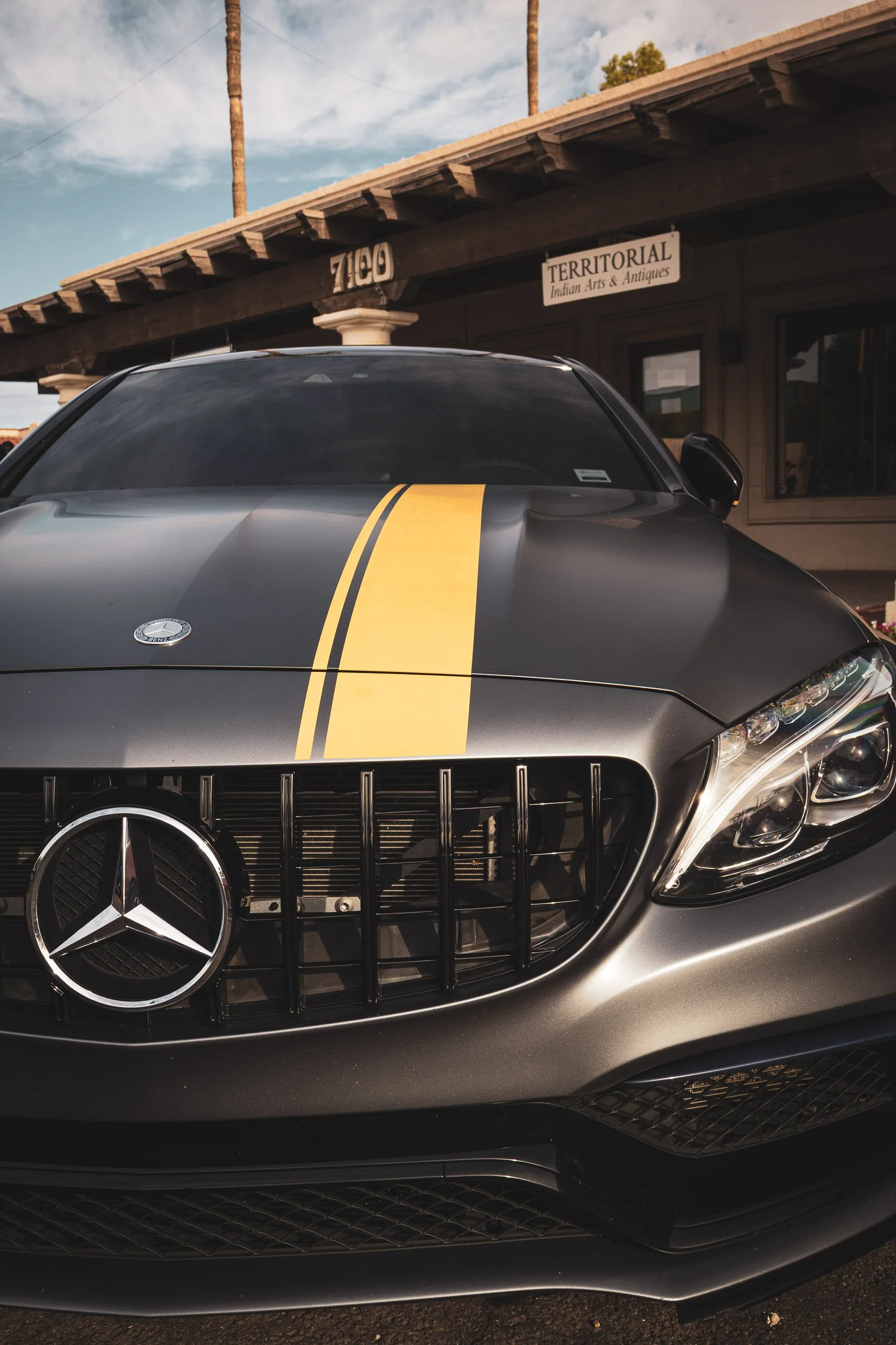 Close-up front automotive photography focusing on the aggressive grille and distinctive yellow hood stripe of a satin gray Mercedes-AMG C63S Edition 1, parked in front of a rustic arts and antiques gallery in Scottsdale, Arizona, car photography by G