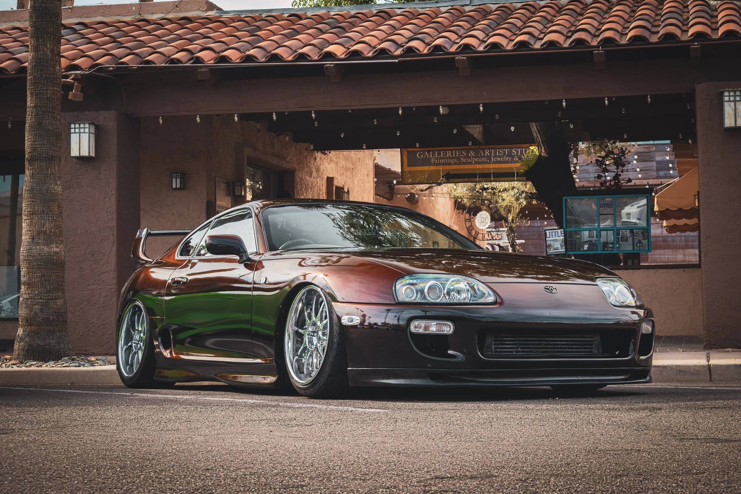 Low-angle front three-quarter automotive photography showcasing a modified, stanced Toyota Supra MK4 featuring a unique colorshift paint finish, deep-dish chrome wheels, and a rear wing, parked on a street in front of an art gallery car meet in Scott