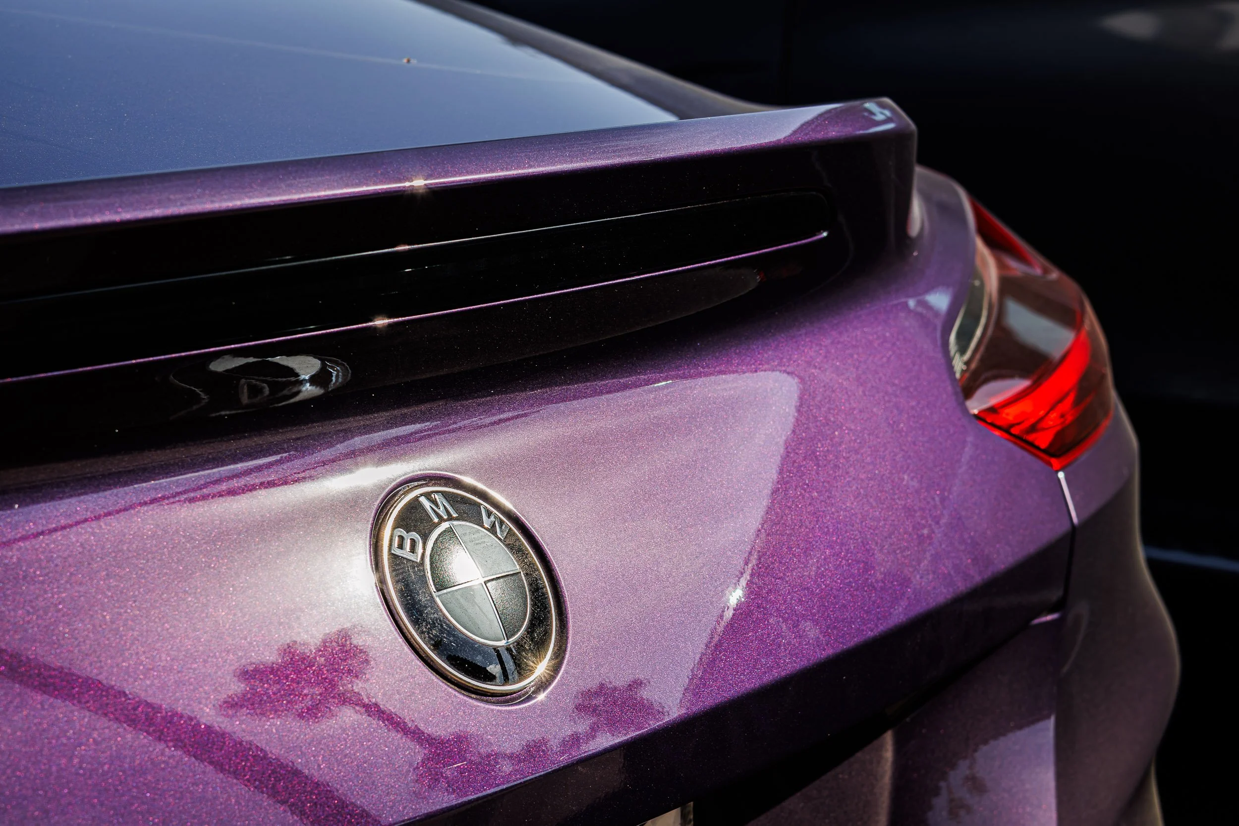 Macro detail automotive photography focusing on the iconic BMW roundel badge and integrated rear lip spoiler, set against vibrant metallic purple paint sparkling in the sun in Chandler, Arizona, captured by Curtis from Gasoline and Dopamine.