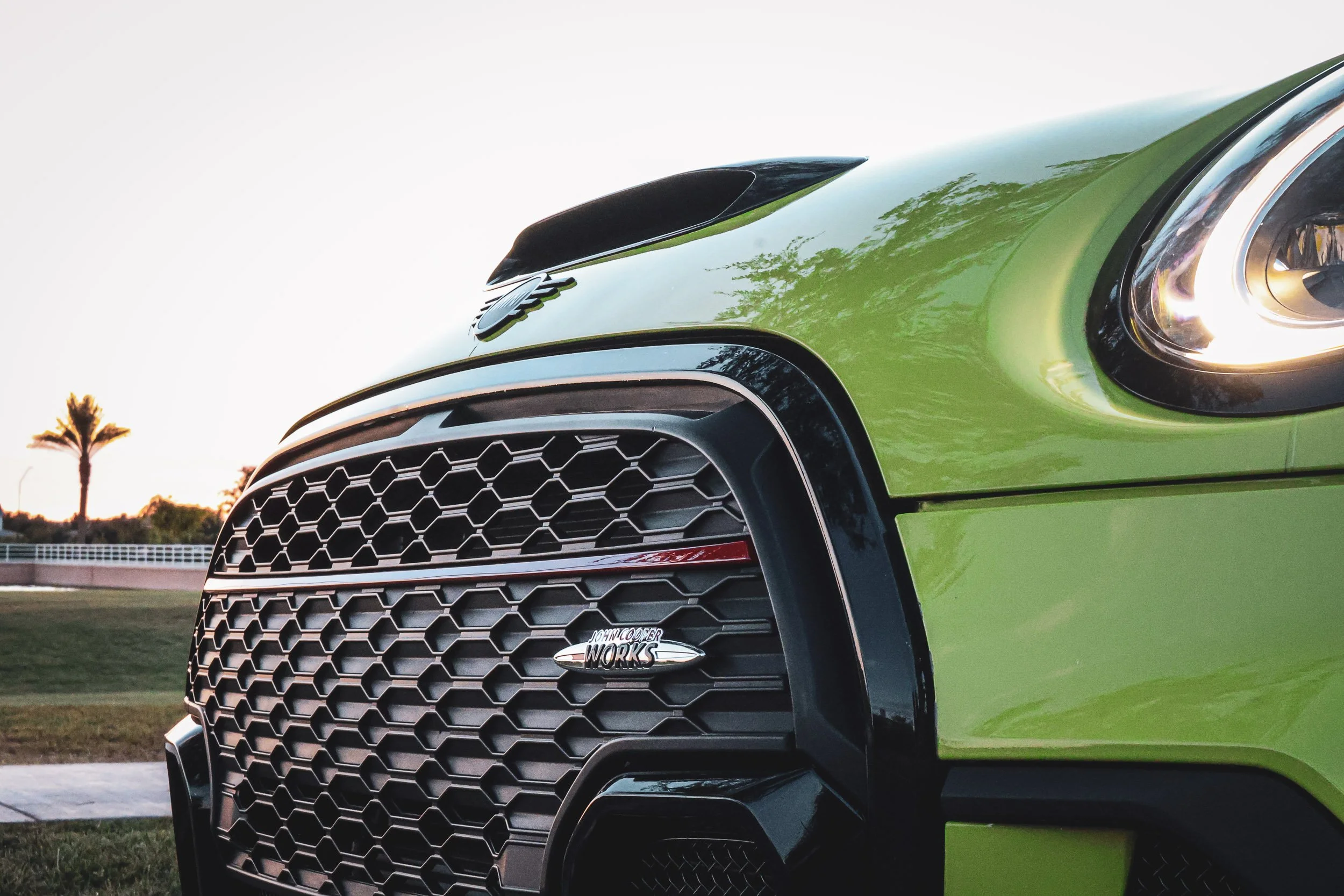 Low-angle macro automotive photography capturing the honeycomb front grille, John Cooper Works badge, and illuminated headlight of a vibrant yellow Mini Cooper JCW, set against a bright sunset sky and a palm tree silhouette in Gilbert, Arizona, captu