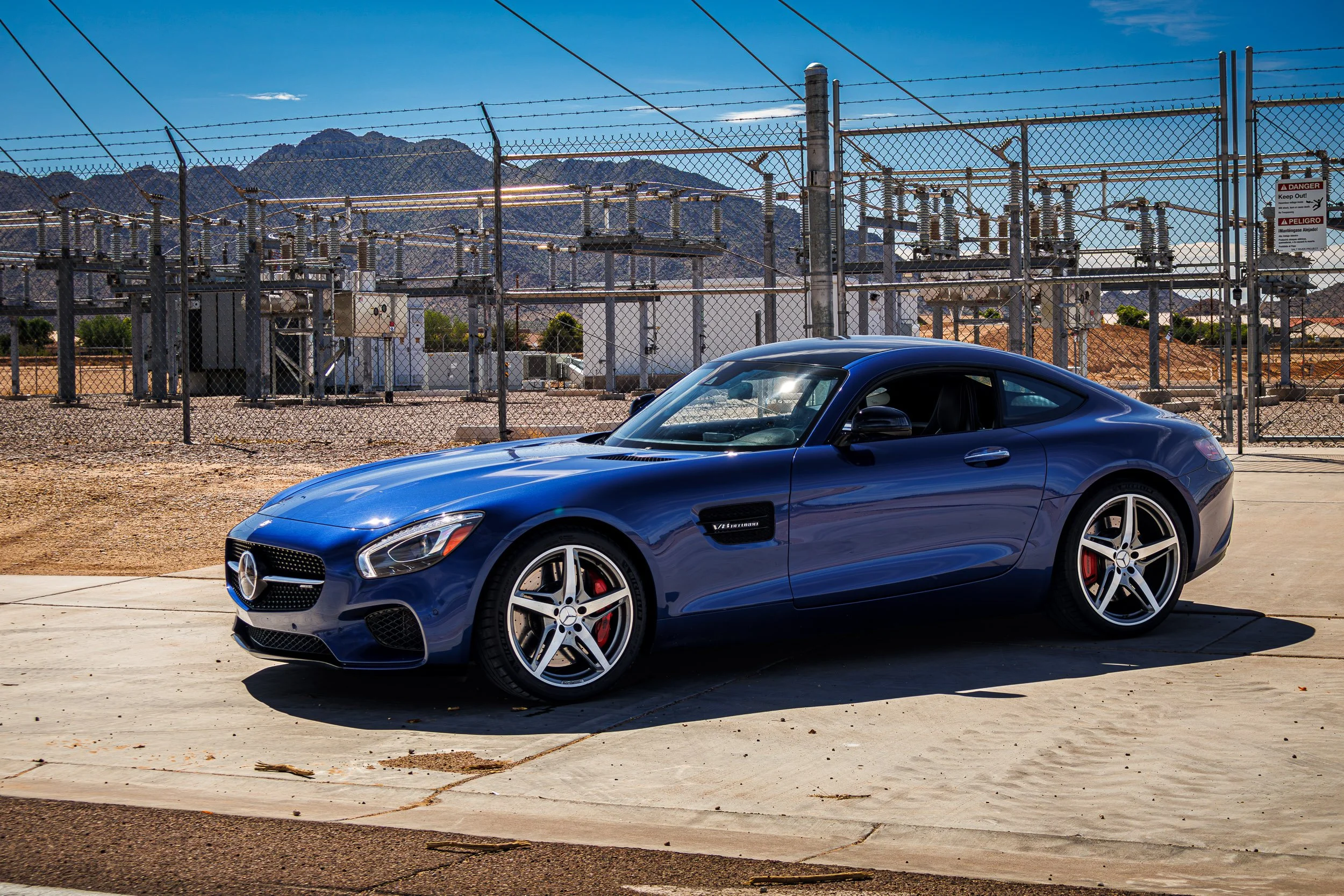 Front three-quarter automotive photography featuring a metallic blue Mercedes-AMG GT S coupe parked in front of an industrial power transformer station, set against a chain-link fence and rugged mountains under a clear blue sky in Queen Creek, Arizon