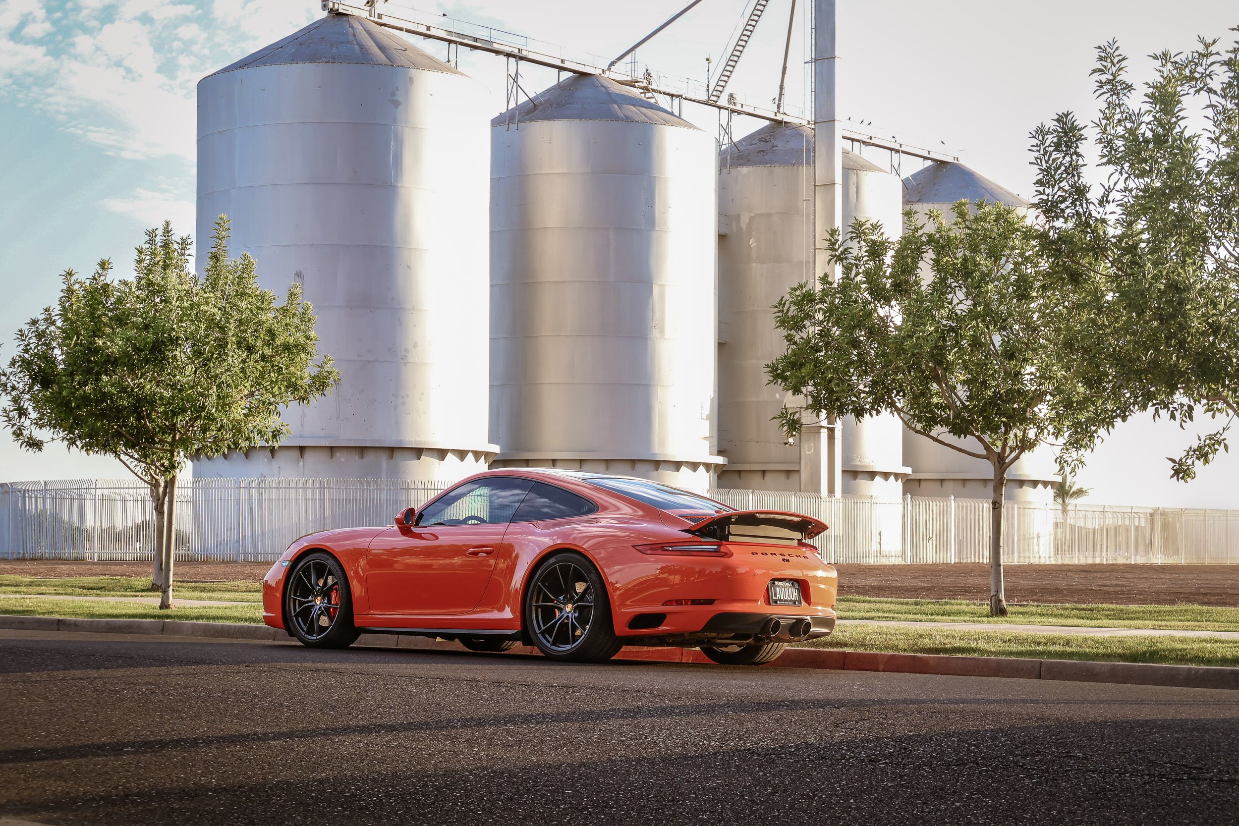 Low-angle rear three-quarter car photography showcasing a Lava Orange Porsche 911 C4S with a raised active spoiler and dual exhaust, parked in front of the historic Morrison Ranch silos in Gilbert, Arizona, captured by Gasoline and Dopamine.