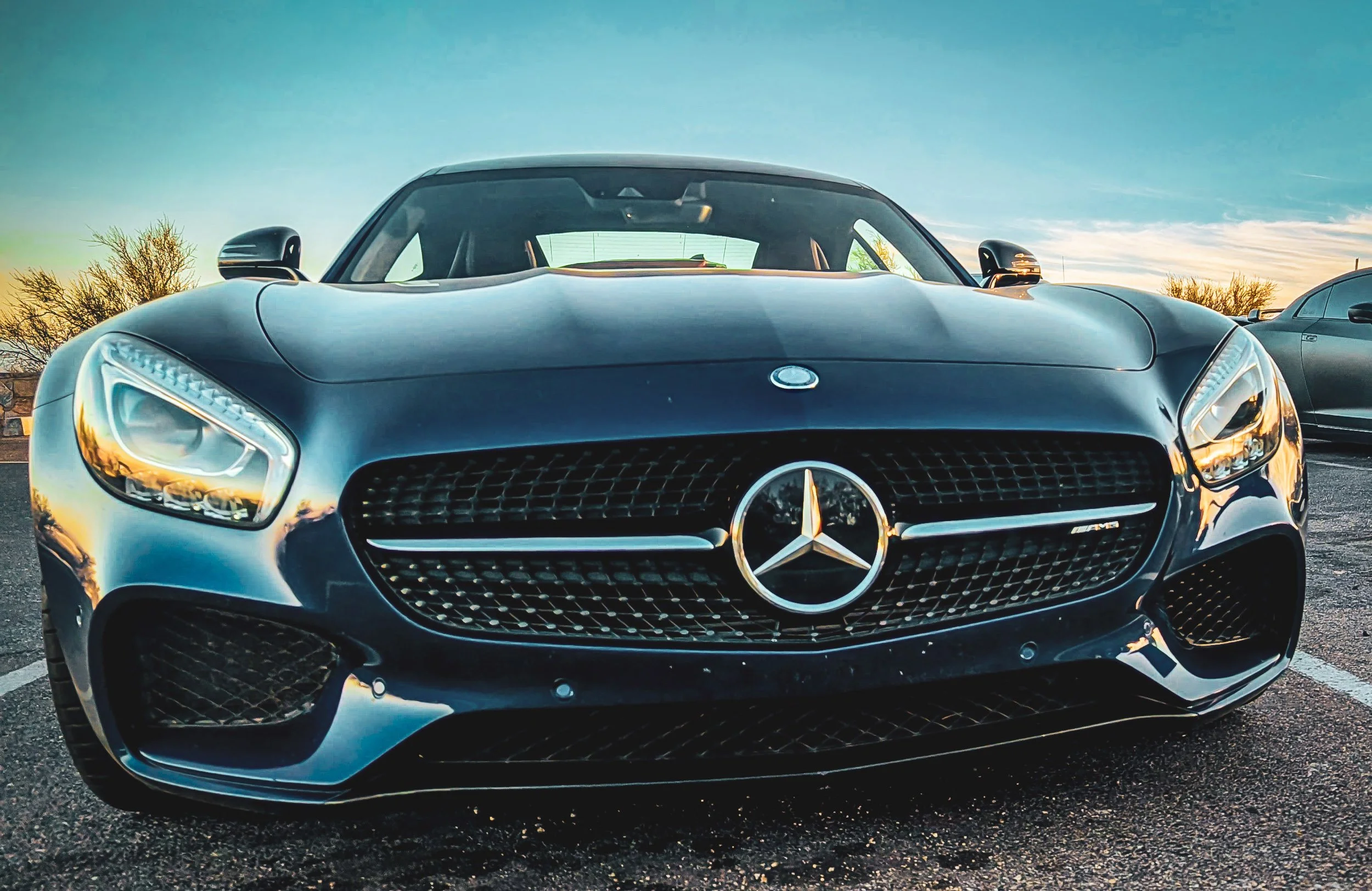 Low-angle front automotive photography highlighting the aggressive front fascia, diamond grille, and sunrise reflections on a metallic blue Mercedes-AMG GT S parked at Dobbins Lookout on South Mountain in Phoenix, Arizona, photographed by Curtis from