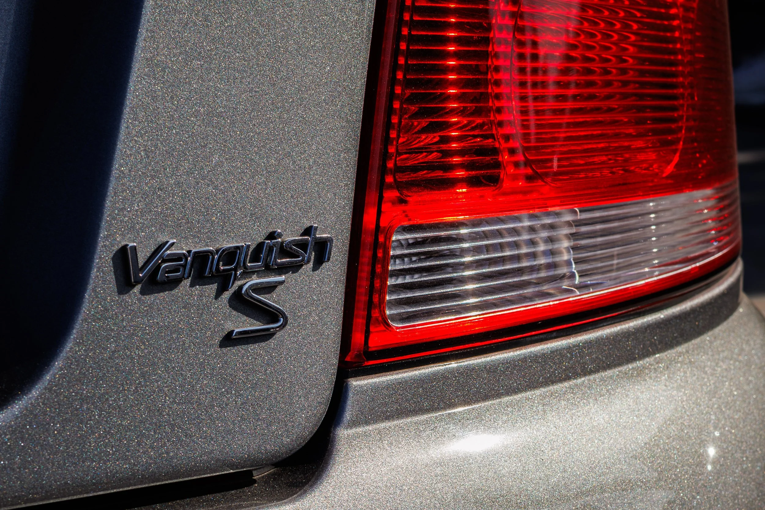 Macro detail automotive photography focusing on the chrome Vanquish S badge and bright red taillight housing set against sparkling silver metallic paint on an Aston Martin in Chandler, Arizona, captured by Curtis from Gasoline and Dopamine.