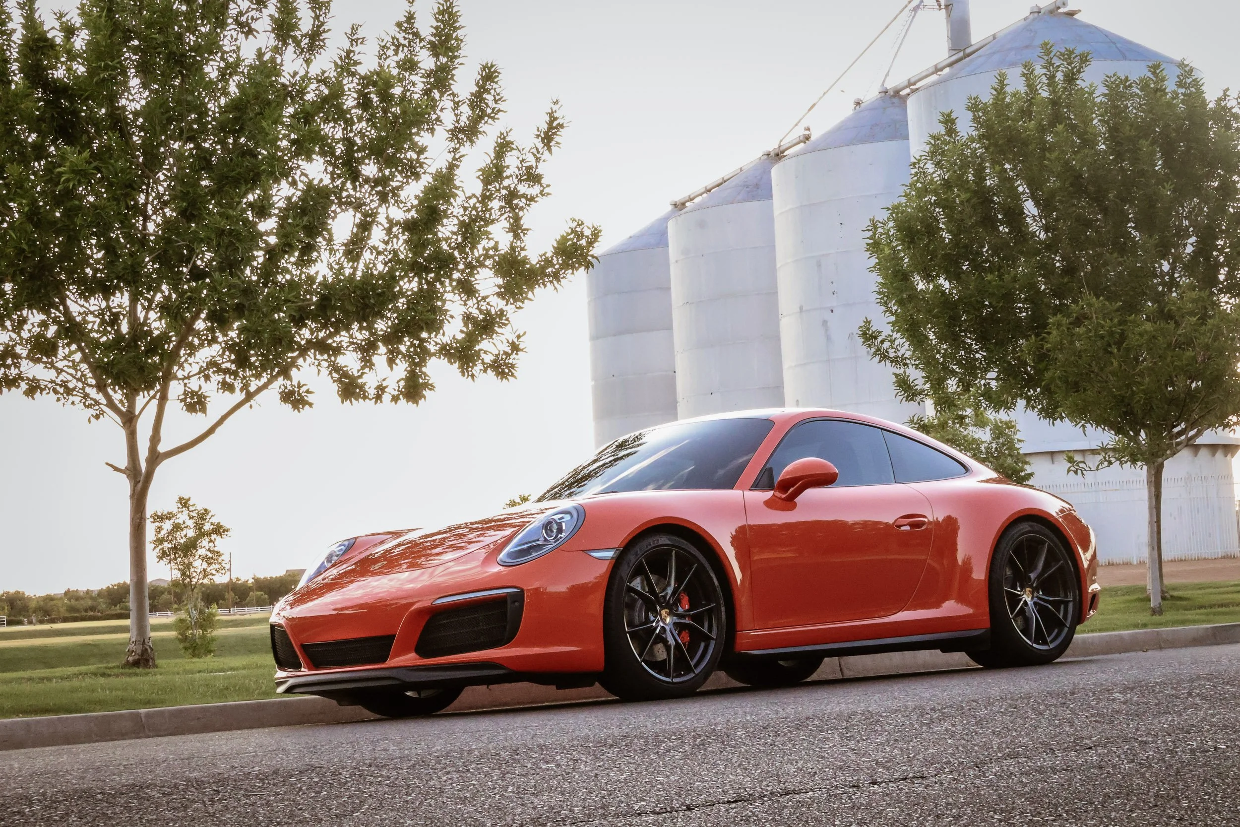 Daytime automotive photography featuring a vibrant Lava Orange Porsche 911 C4S parked in front of the historic Morrison Ranch silos in Gilbert, Arizona, captured by Gasoline and Dopamine.