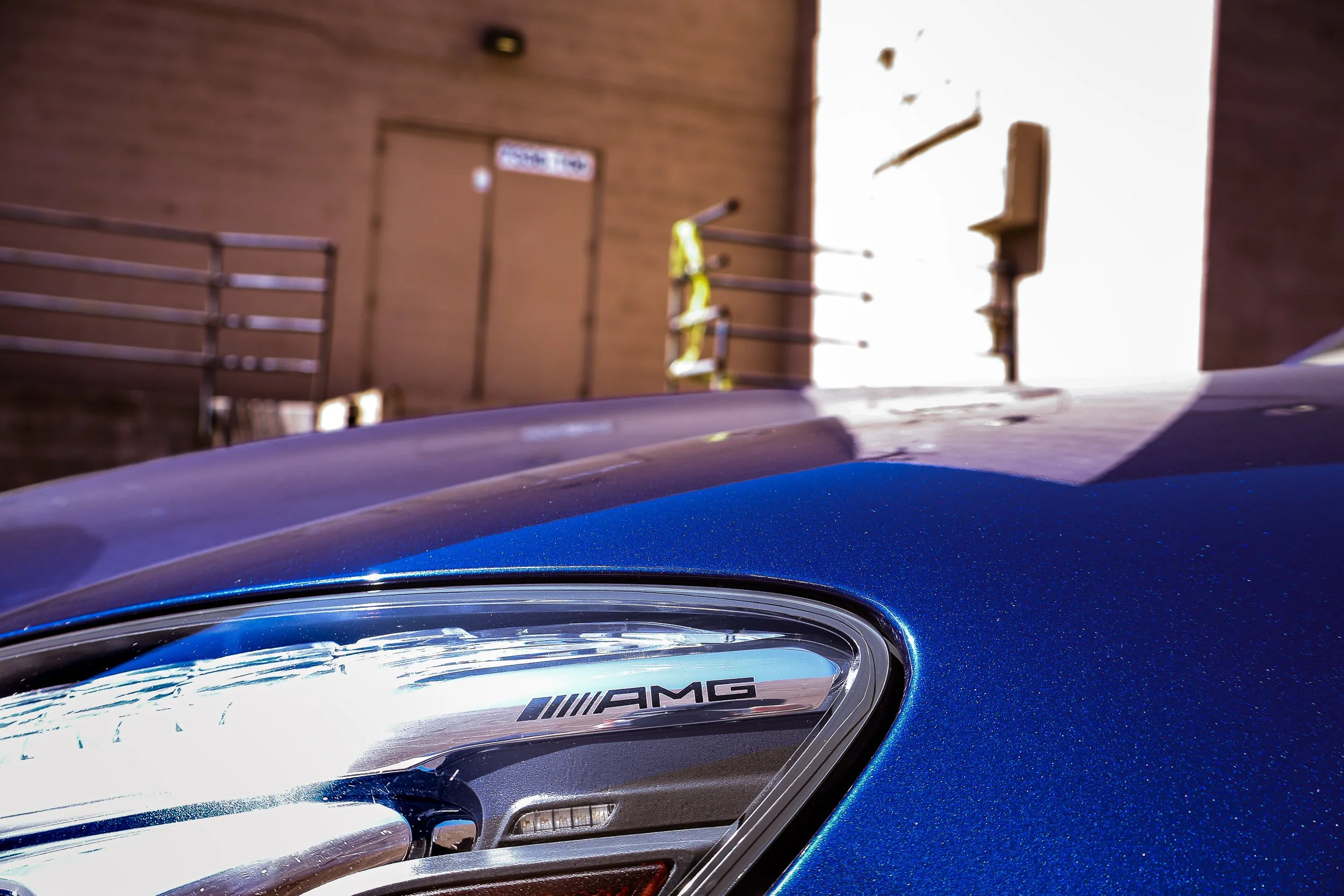 Macro automotive detail photography focusing on the etched AMG logo inside the headlight housing and the sparkling metallic blue paint of a Mercedes-AMG GT S, set against a blurred industrial alleyway under harsh midday sunlight in Queen Creek, Arizo