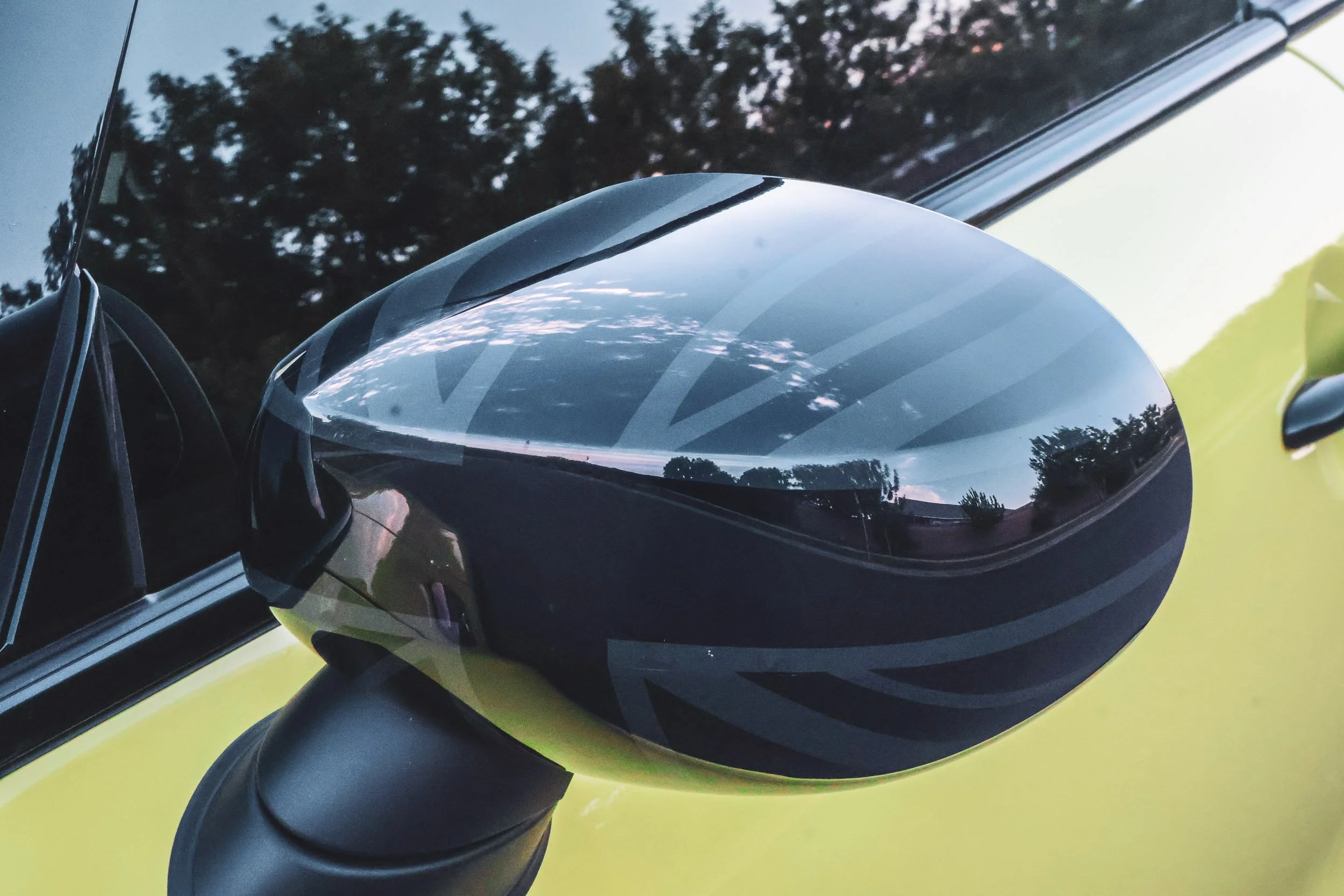 Macro automotive detail photography highlighting a subtle dark Union Jack graphic on the side mirror of a yellow Mini Cooper JCW, reflecting the dusk sky and neighborhood trees in Gilbert, Arizona, shot by Curtis at Gasoline and Dopamine.