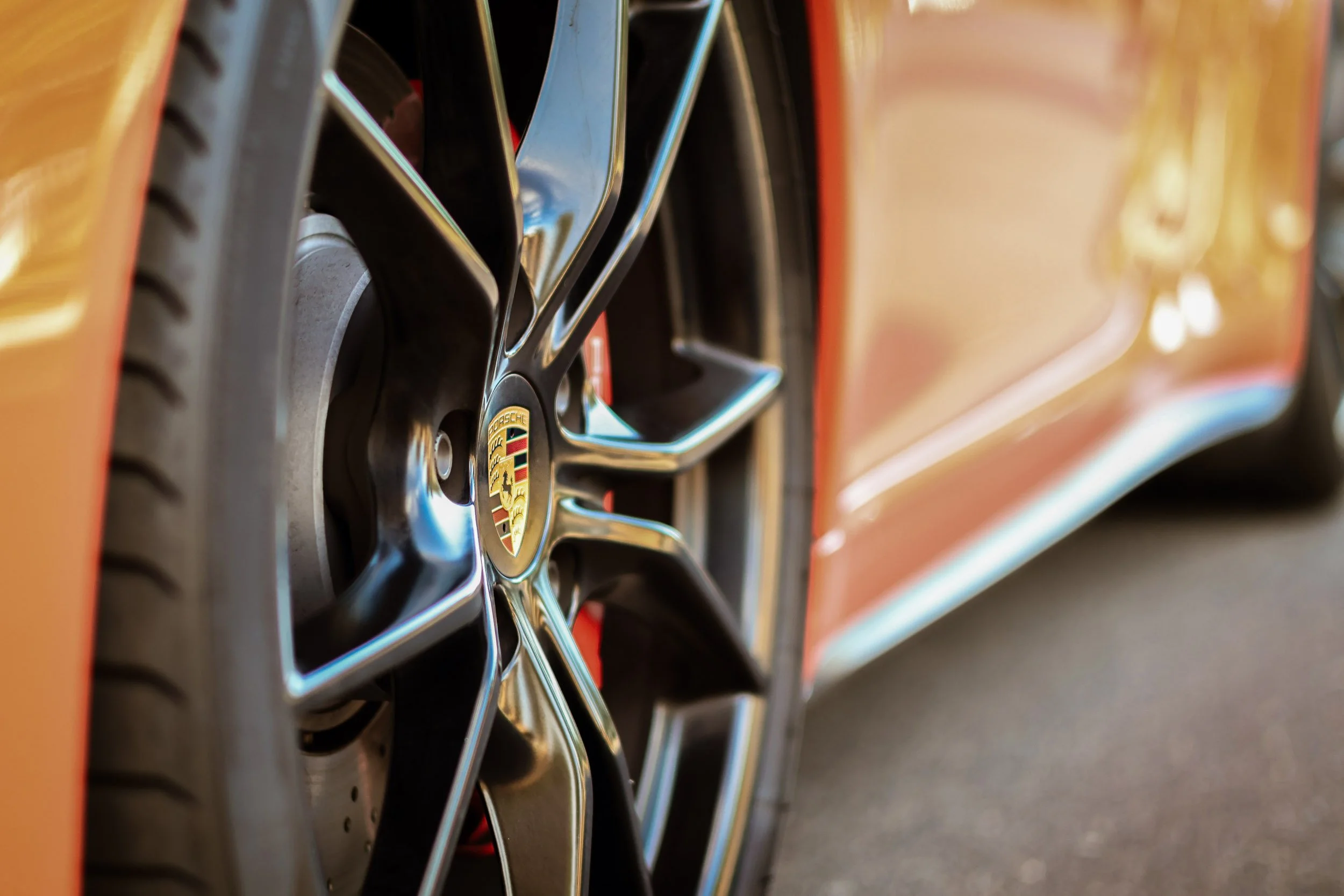 Macro detail car photography focusing on the iconic Porsche center cap crest and dark multi-spoke wheel, set against the blurred Lava Orange bodywork of a 911 C4S in Gilbert, Arizona, captured by Curtis from Gasoline and Dopamine.