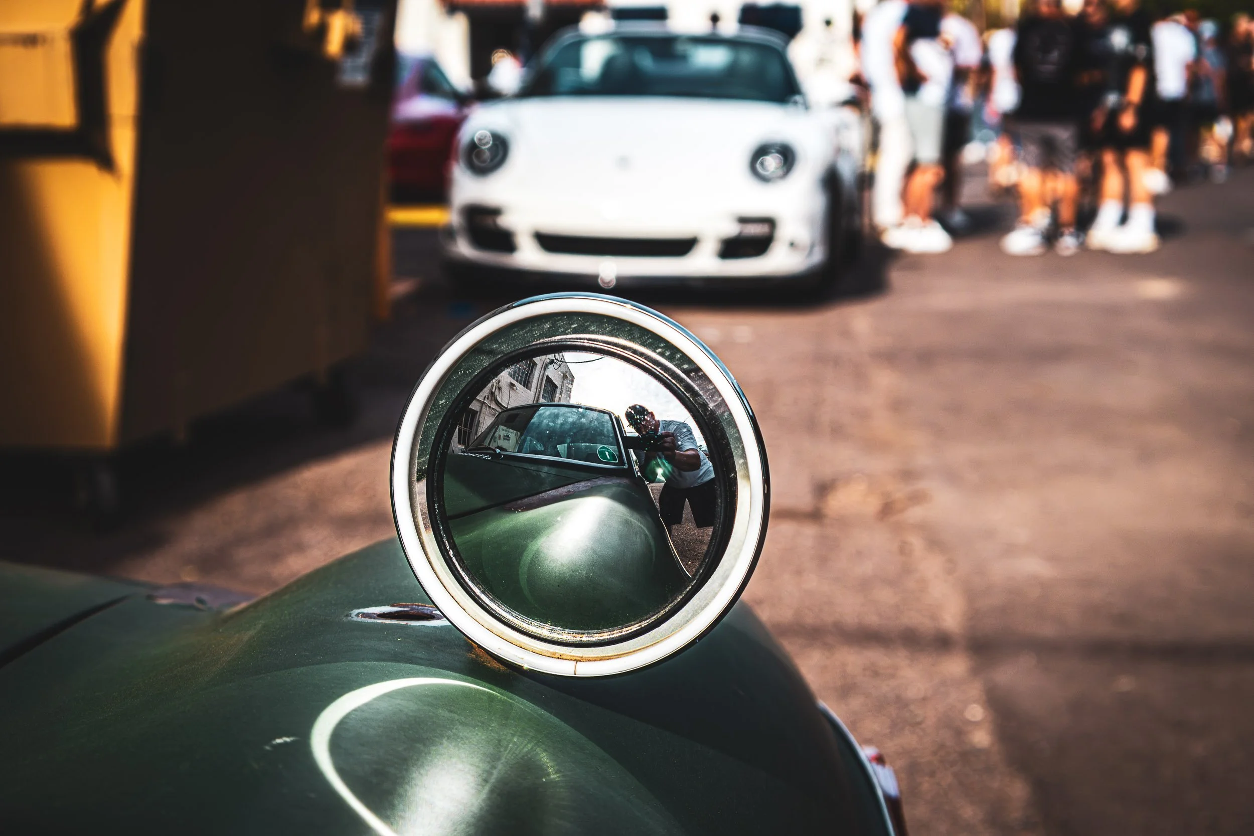 Macro automotive detail photography focusing on the round chrome fender mirror of a classic dark green MG B GT, capturing a self-portrait reflection of the photographer, Curtis, against the blurred backdrop of a lively cars and coffee event and a whi