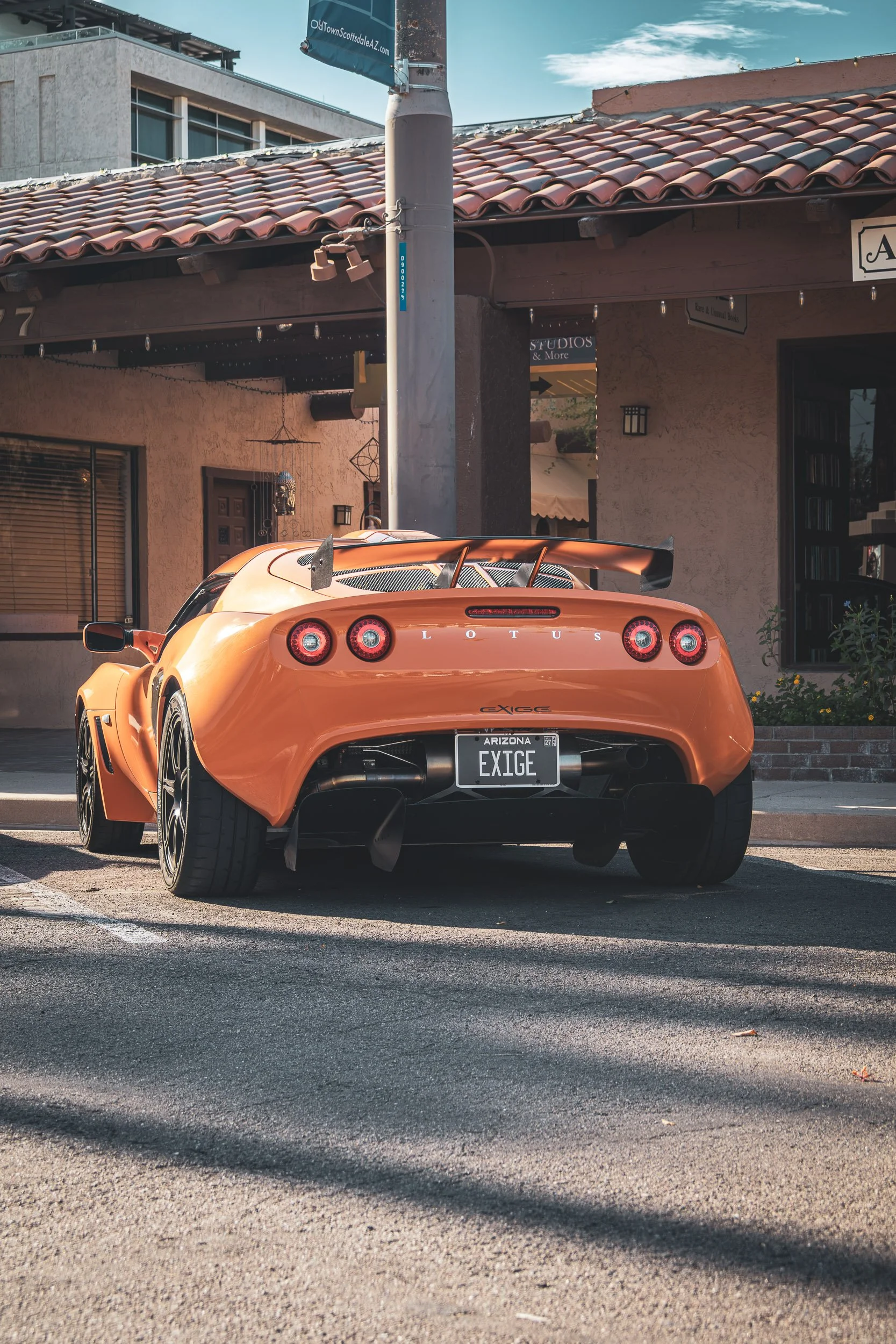 Low-angle rear three-quarter automotive photography featuring a vibrant orange Lotus Exige parked on a sunny street in front of adobe storefronts in Old Town Scottsdale, Arizona, captured by Curtis at Gasoline and Dopamine.