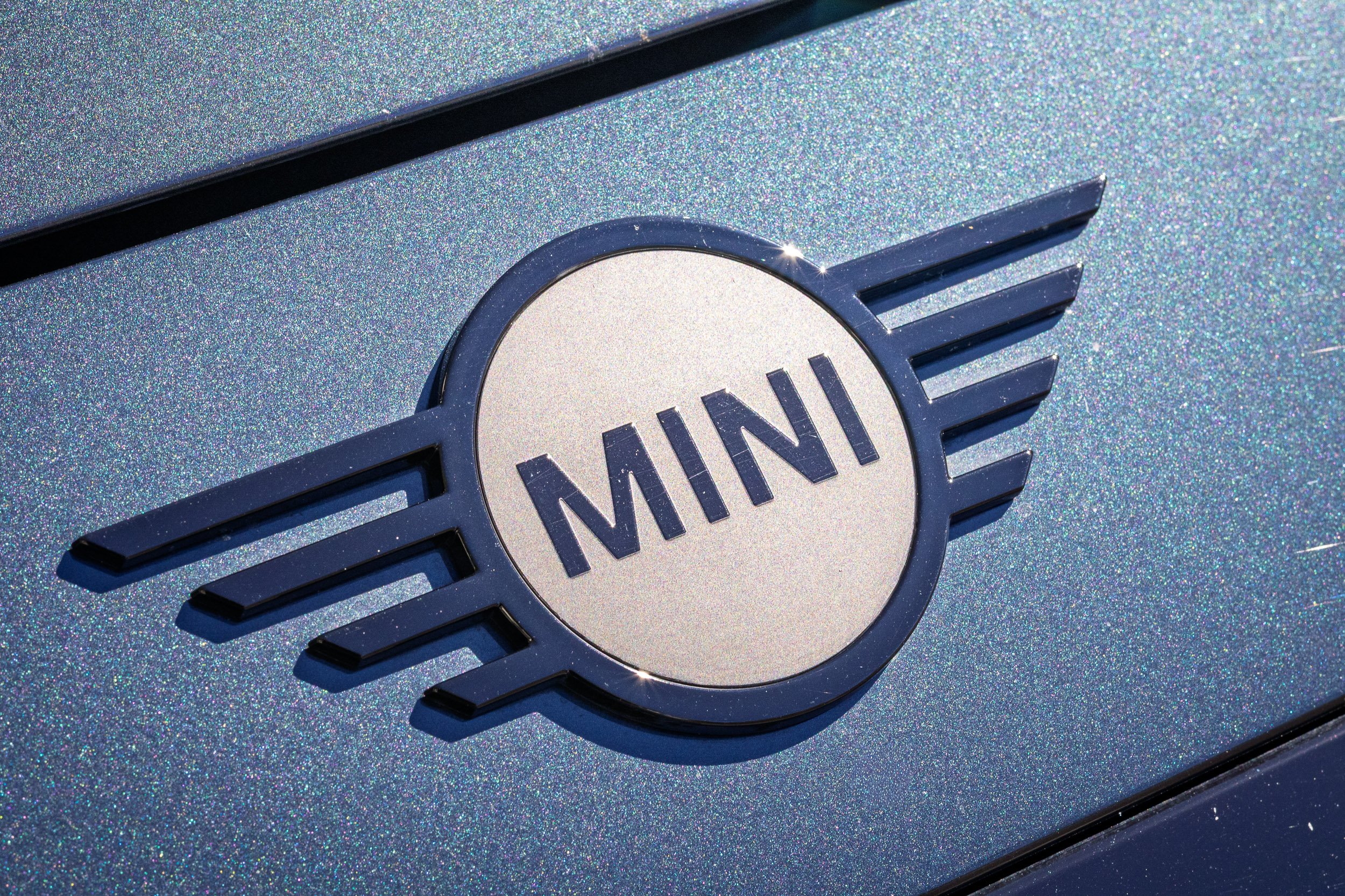 Macro detail car photography focusing on a black and silver MINI emblem set against a vibrant metallic blue paint flake in Tempe, Arizona, photographed by Gasoline and Dopamine.