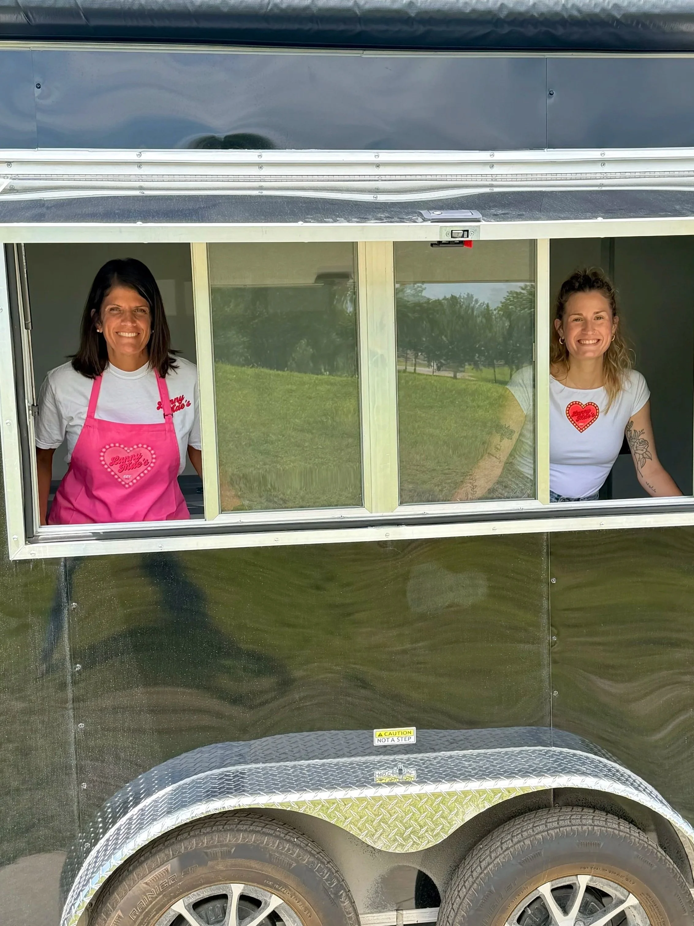 A black food truck with two women inside, one wearing a pink apron and the other wearing a white t-shirt with a heart on it. They are smiling and looking out of the windows.