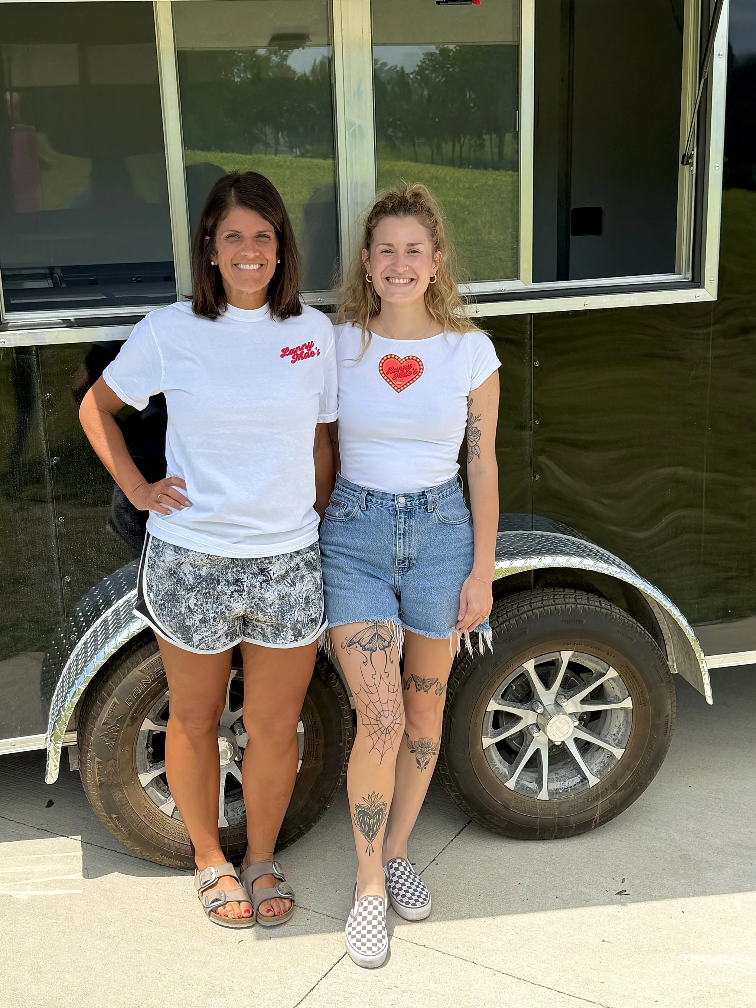 Two women standing in front of a black trailer with a window, smiling. The woman on the left has dark hair, wearing a white T-shirt with red text and patterned shorts. The woman on the right has blonde hair, tattoos, wearing a white T-shirt with red and yellow heart graphic, and denim shorts.