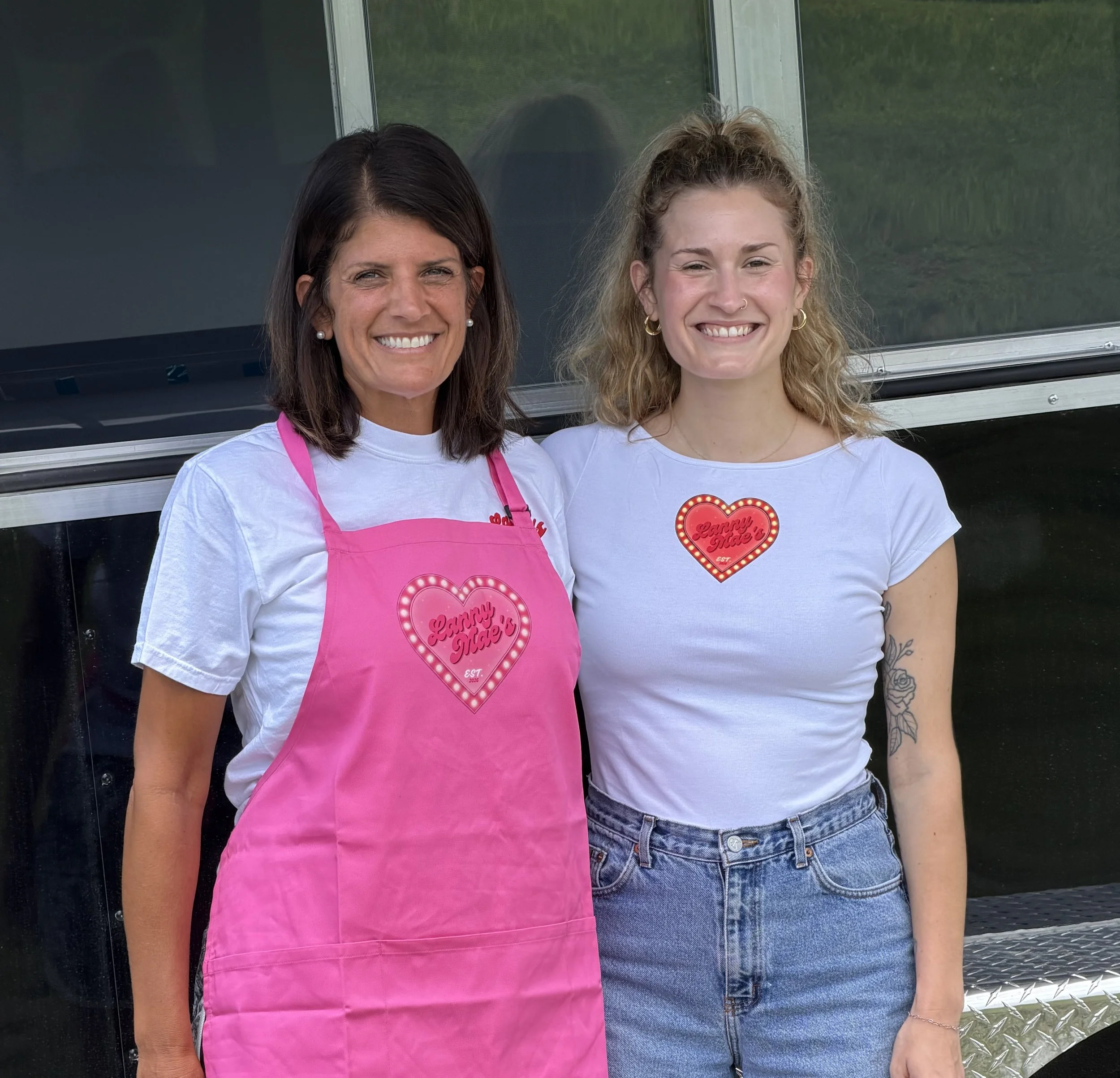 Two women standing side by side outdoors, smiling at the camera. The woman on the left has dark hair, earrings, and is wearing a white shirt with a pink apron that says "Lanny Mae's" inside a heart shape. The woman on the right has curly blonde hair, a nose ring, earrings, a white t-shirt with a heart-shaped logo, and a tattoo on her right forearm. They are in front of a green grass background and a black and silver structure.