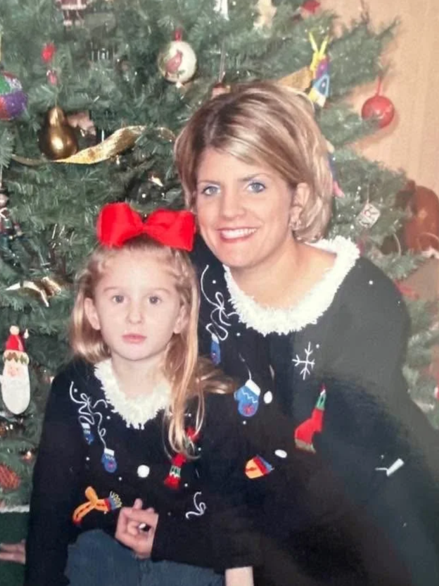 A woman and a young girl pose in front of a decorated Christmas tree, both wearing matching Christmas sweaters.