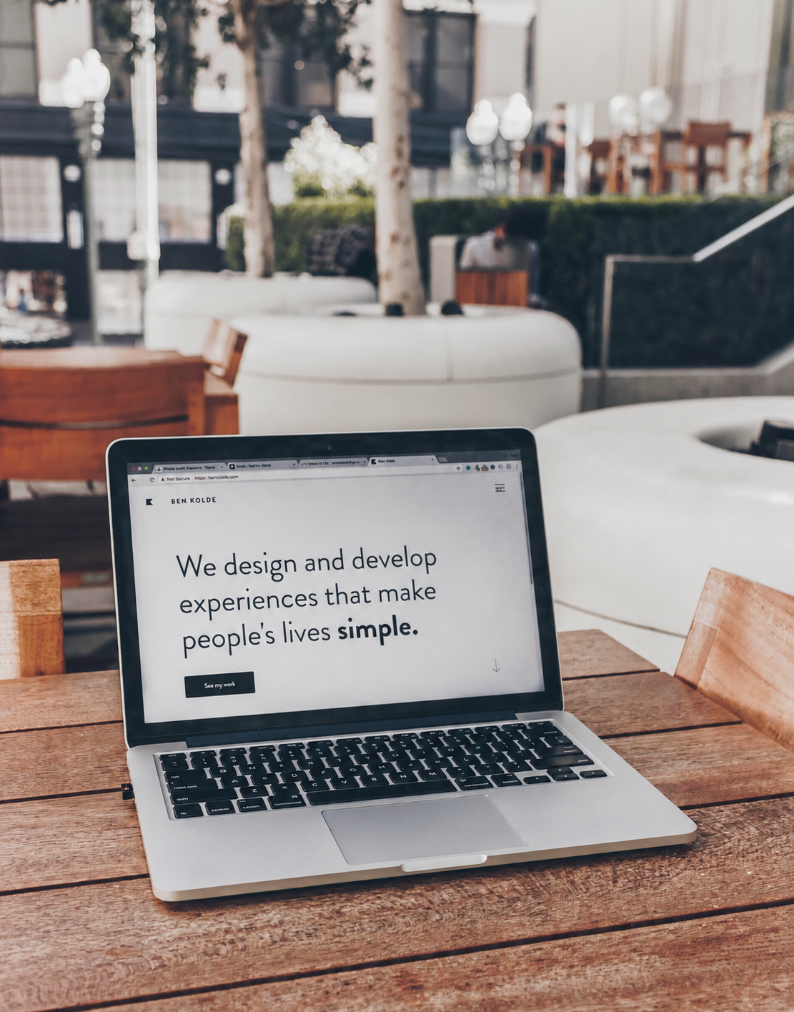 A laptop on a wooden table displaying a website with the text: "We design and develop experiences that make people's lives simple." The setting appears to be an outdoor cafe or patio with trees, chairs, and lampposts in the background.