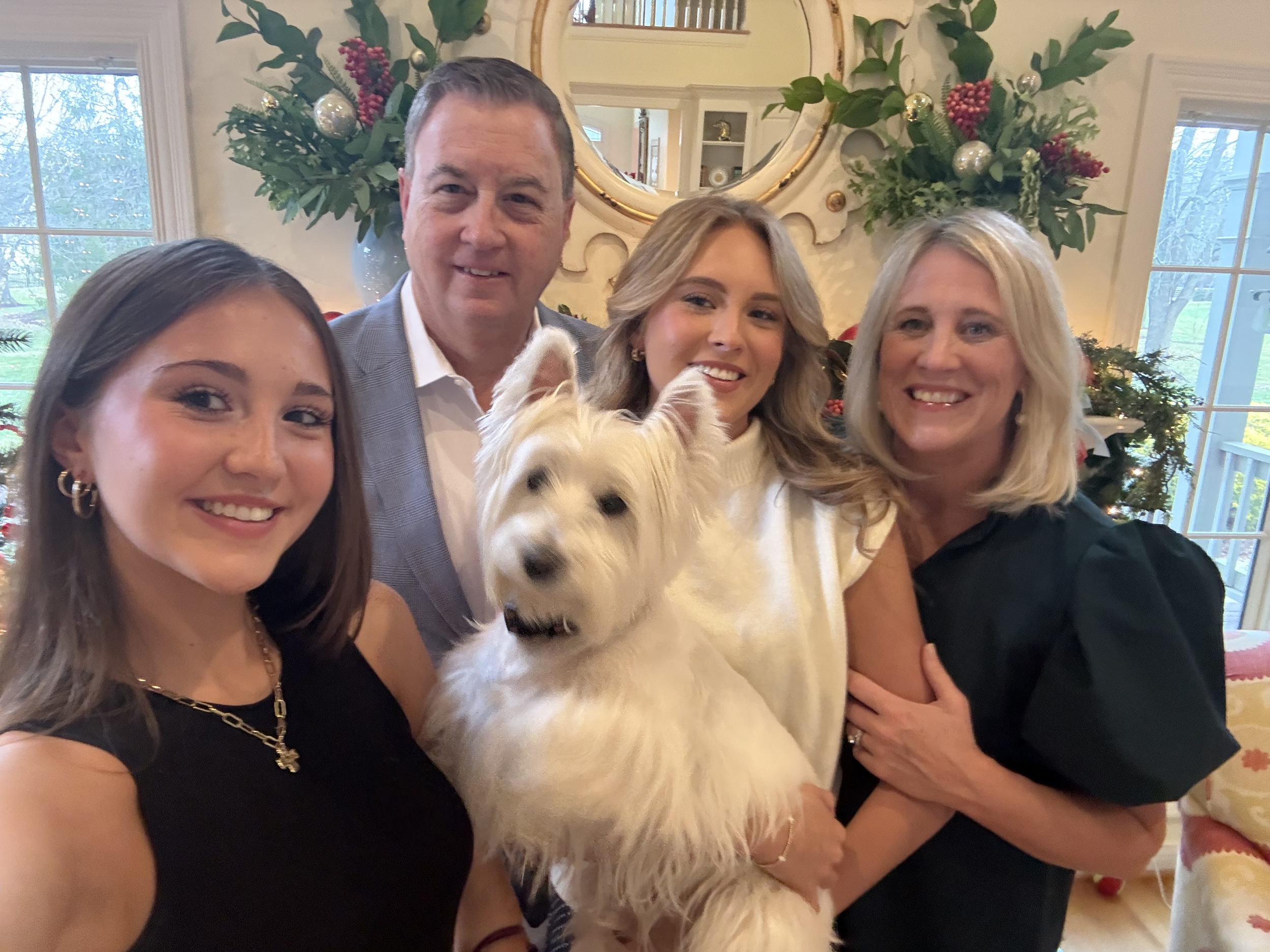 Group of five people, including a dog, celebrating in a decorated room with windows and festive holiday decorations.