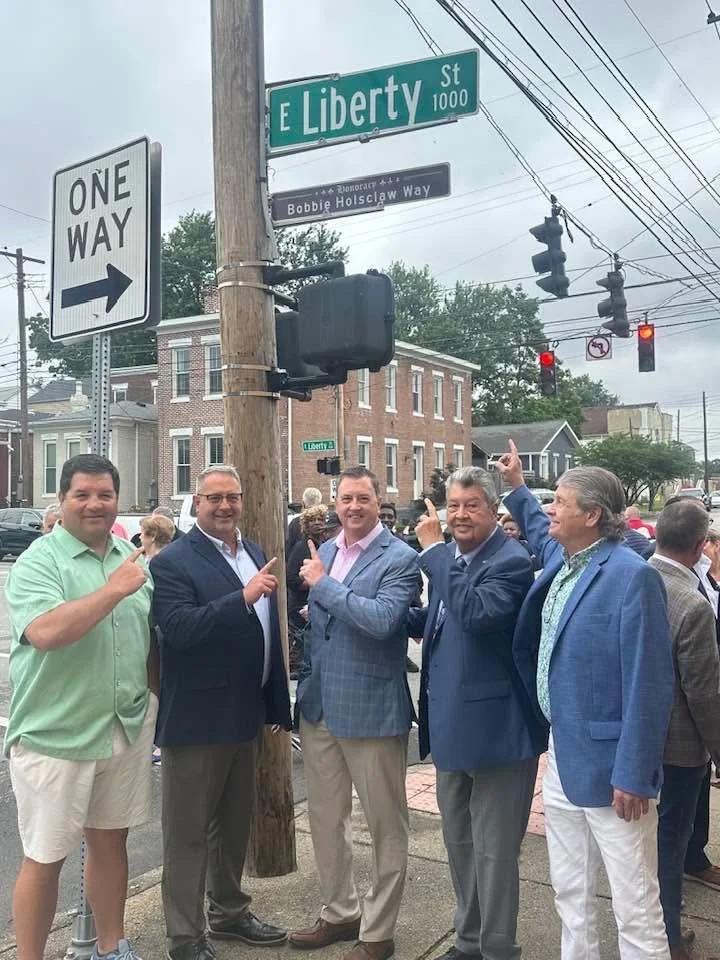 Group of men at a street corner pointing at a street sign that reads 'E Liberty St' at an intersection with traffic lights and other street signs, during a cloudy day.
