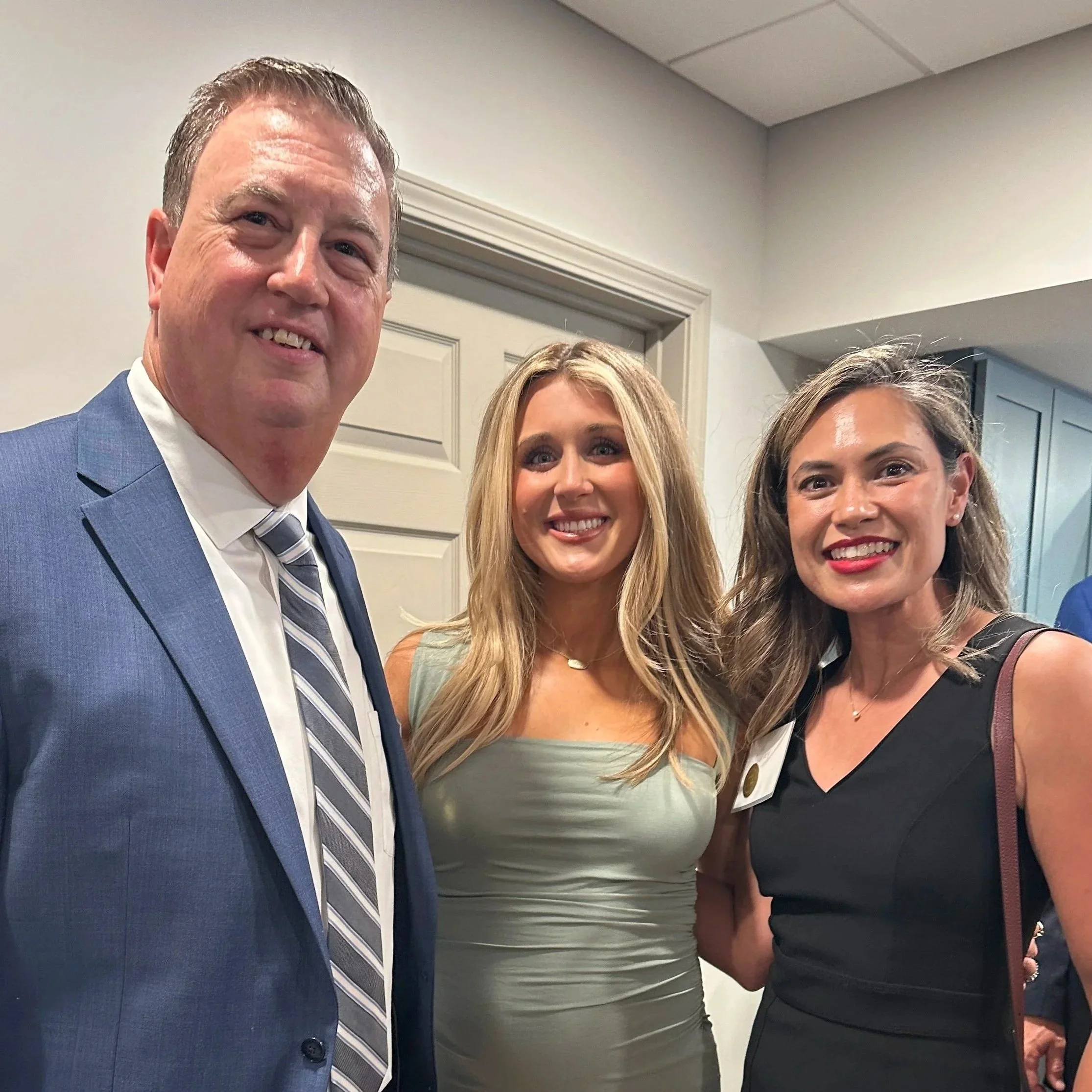 Close-up of three people posing for a photo indoors, with part of a man in a blue suit and two women visible, one with blonde hair and the other with brown hair.