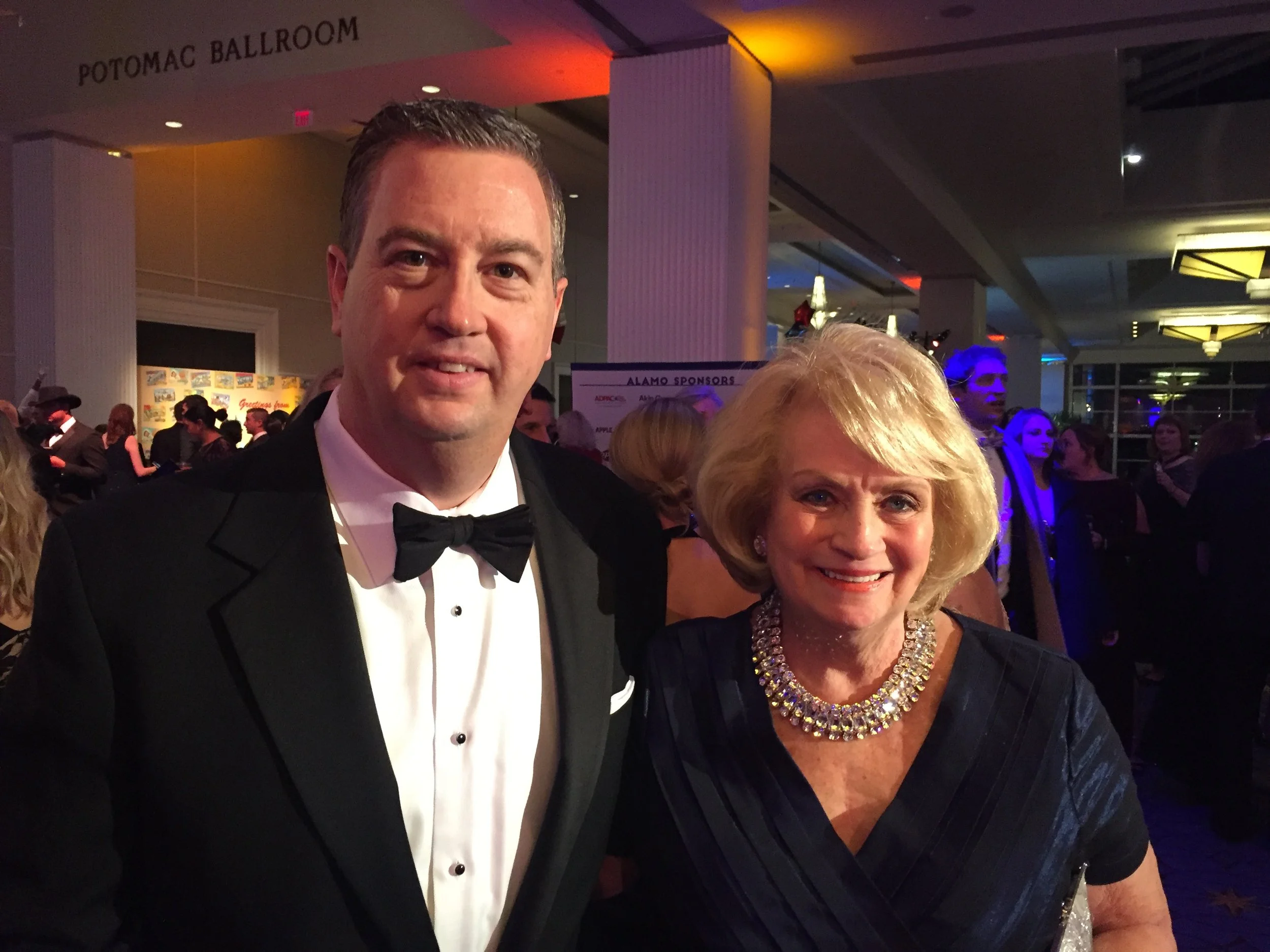 A man in a tuxedo and a woman in a dark dress with a bejeweled necklace posing at a formal event in a ballroom.